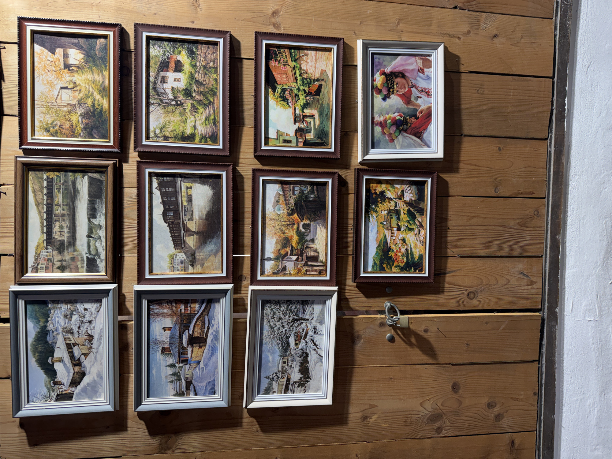 A collection of framed pictures hangs on a light brown wooden wall with horizontal planks, likely inside a building in Lovech, Bulgaria. The wall has a latch on it. The pictures vary in size, shape, and frame color (brown and white). The paintings depict various scenes, including landscapes with houses, bridges, and winter scenes. One picture includes a person wearing traditional clothing. The right side of the image shows a white wall adjacent to the wooden planks. The pictures are arranged in rows and columns, suggesting a deliberate arrangement on the wall.