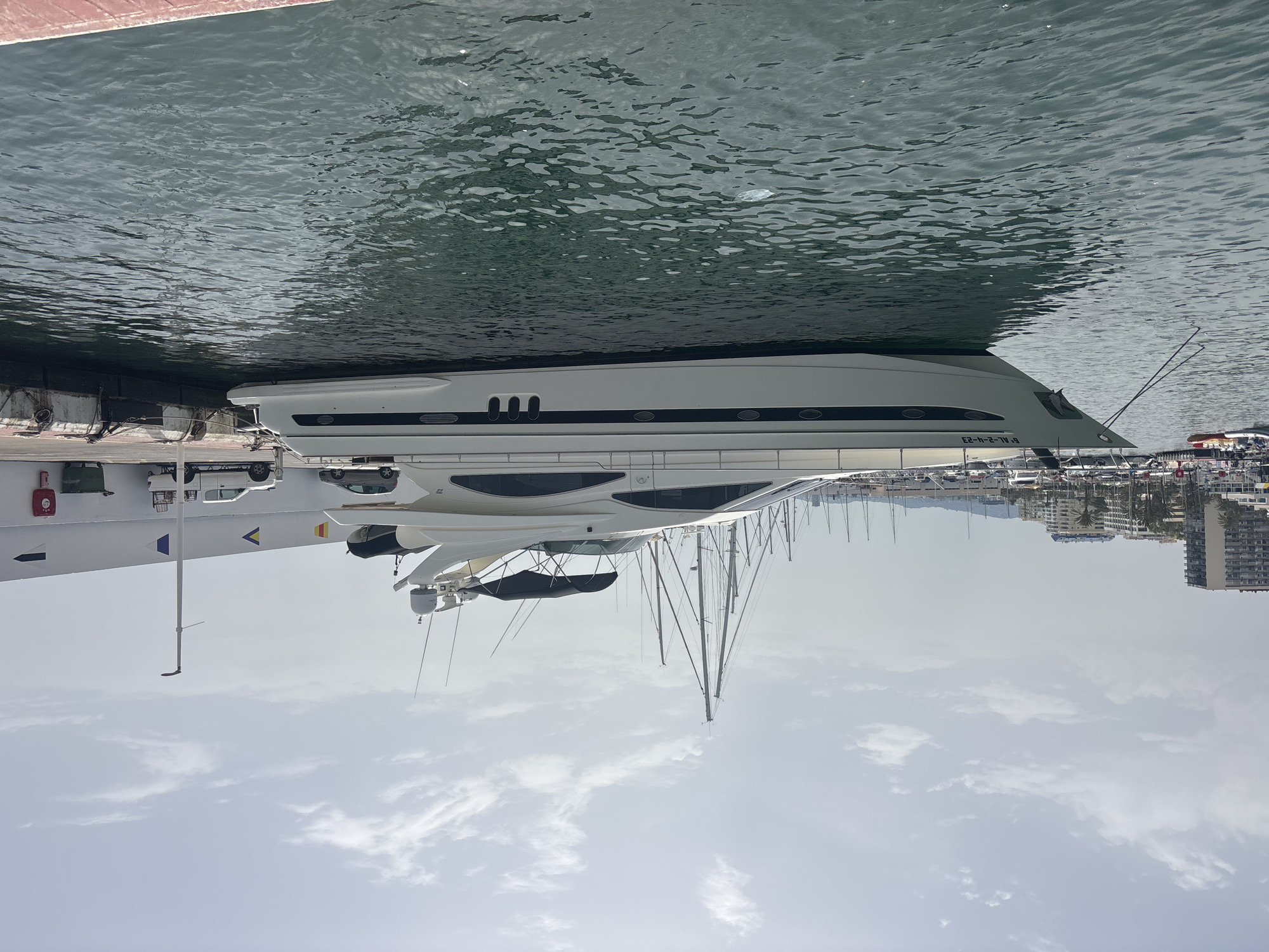 The image shows a large white yacht docked in a marina in Marbella, Spain. The yacht is positioned parallel to a concrete pier. Behind the yacht, other boats with masts are visible, and further in the background, buildings and palm trees line the shore. On the pier, a white pickup truck and a silver car are parked. The sky is partly cloudy. The water in the foreground is a dark green color with visible ripples. The pier has a white wall with several colored flags and a fire extinguisher mounted on it.