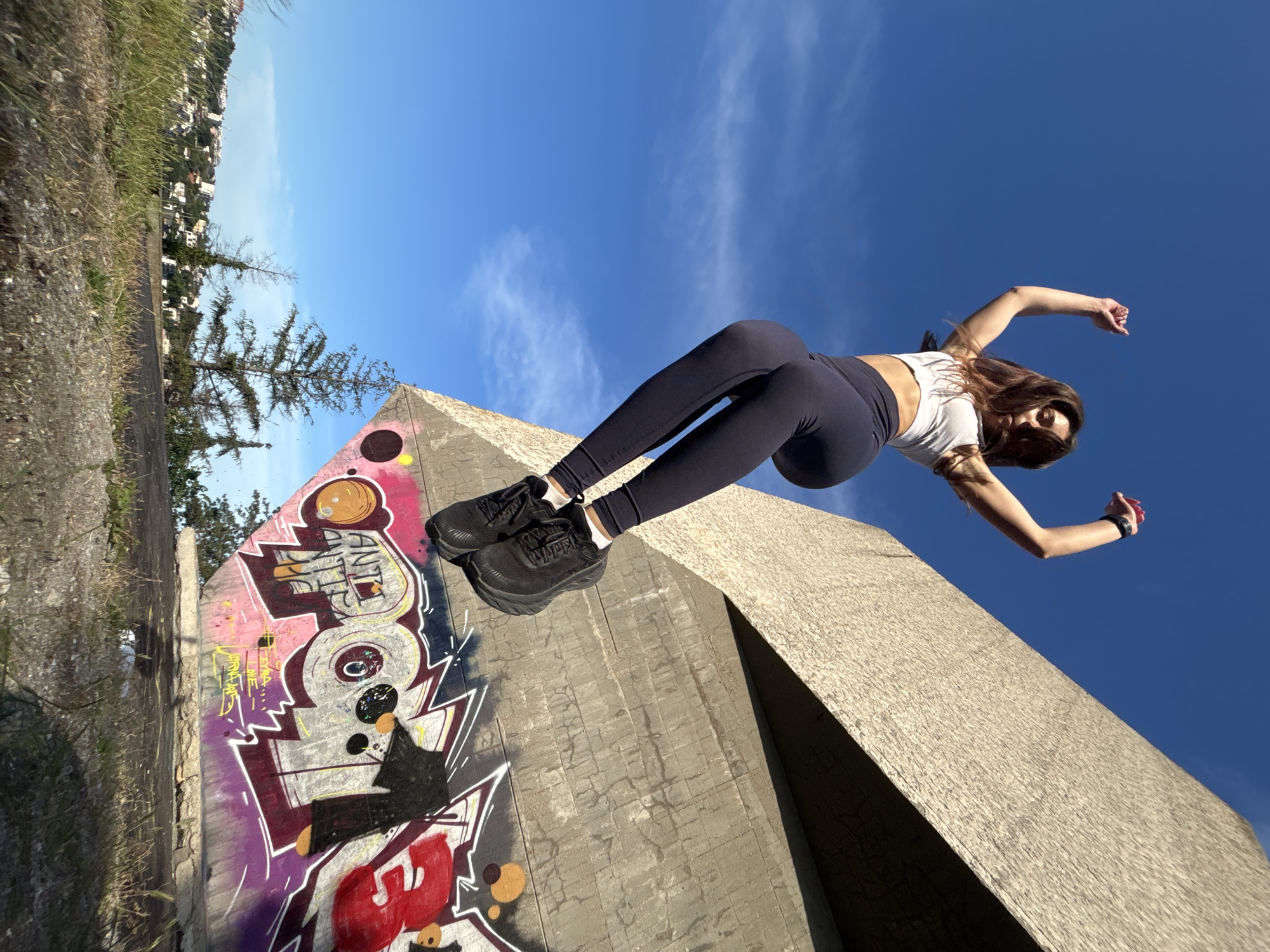 A young woman with long brown hair is captured mid-air, jumping in front of a concrete structure adorned with graffiti. She is wearing a white sports bra, dark leggings, and black athletic shoes. Her arms are raised, and her legs are bent, suggesting an energetic movement. The concrete structure is angular and textured, with vibrant graffiti tags covering its lower section. Behind the structure, a clear blue sky dominates the upper portion of the image, with a few wispy clouds visible. To the left and in the background, a cityscape with buildings is nestled amongst trees on a hill, characteristic of Varna, Bulgaria. The foreground consists of dry grass and rocky ground.