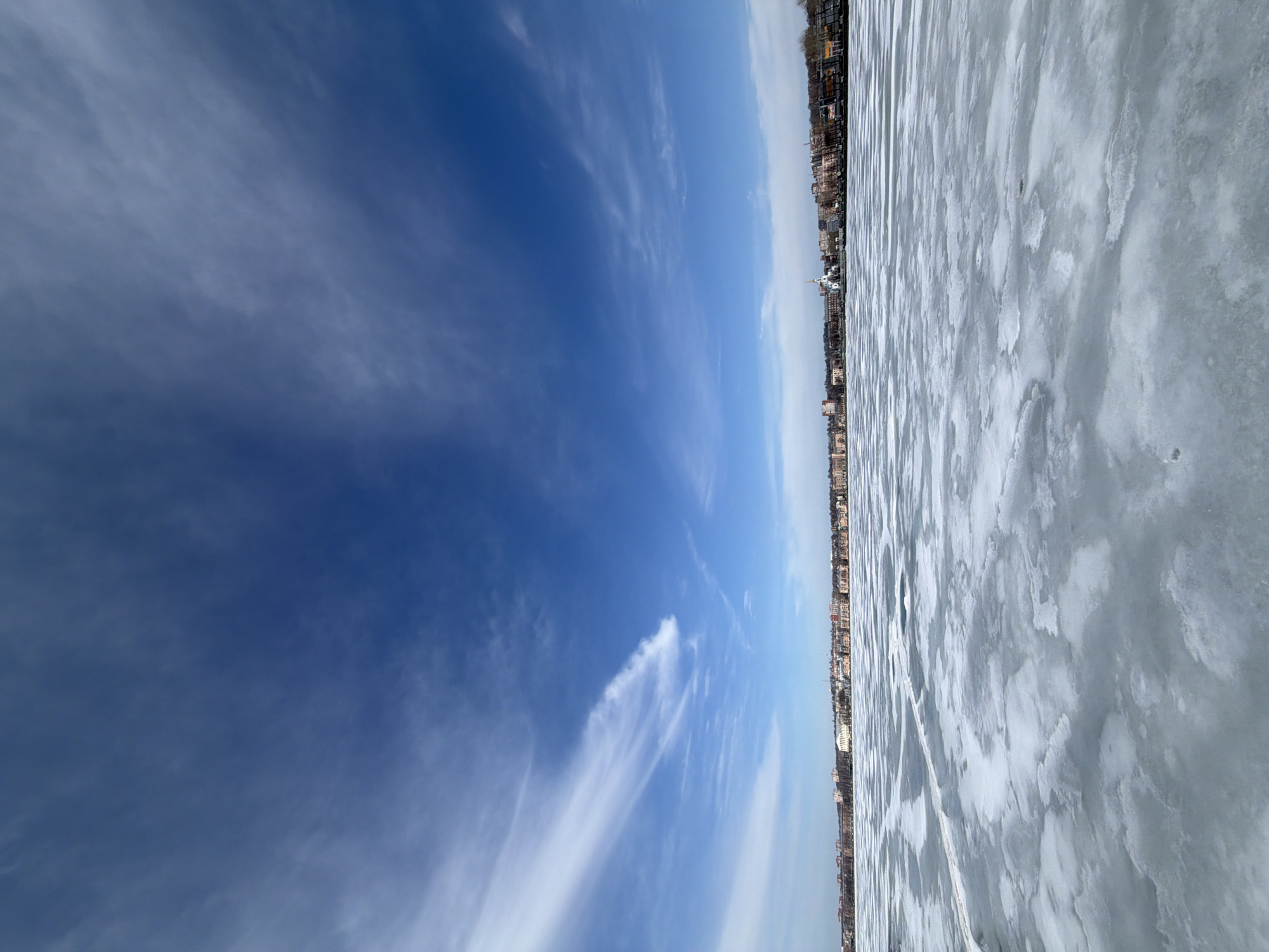 The image shows a winter scene in Votkinsk, Russia. The right side of the image depicts a frozen body of water, possibly the Votkinsk Reservoir, with a cracked ice surface. On the horizon is the cityscape of Votkinsk, with buildings and possibly a church steeple visible. The left side of the image shows a clear, blue sky with streaks of cirrus clouds.