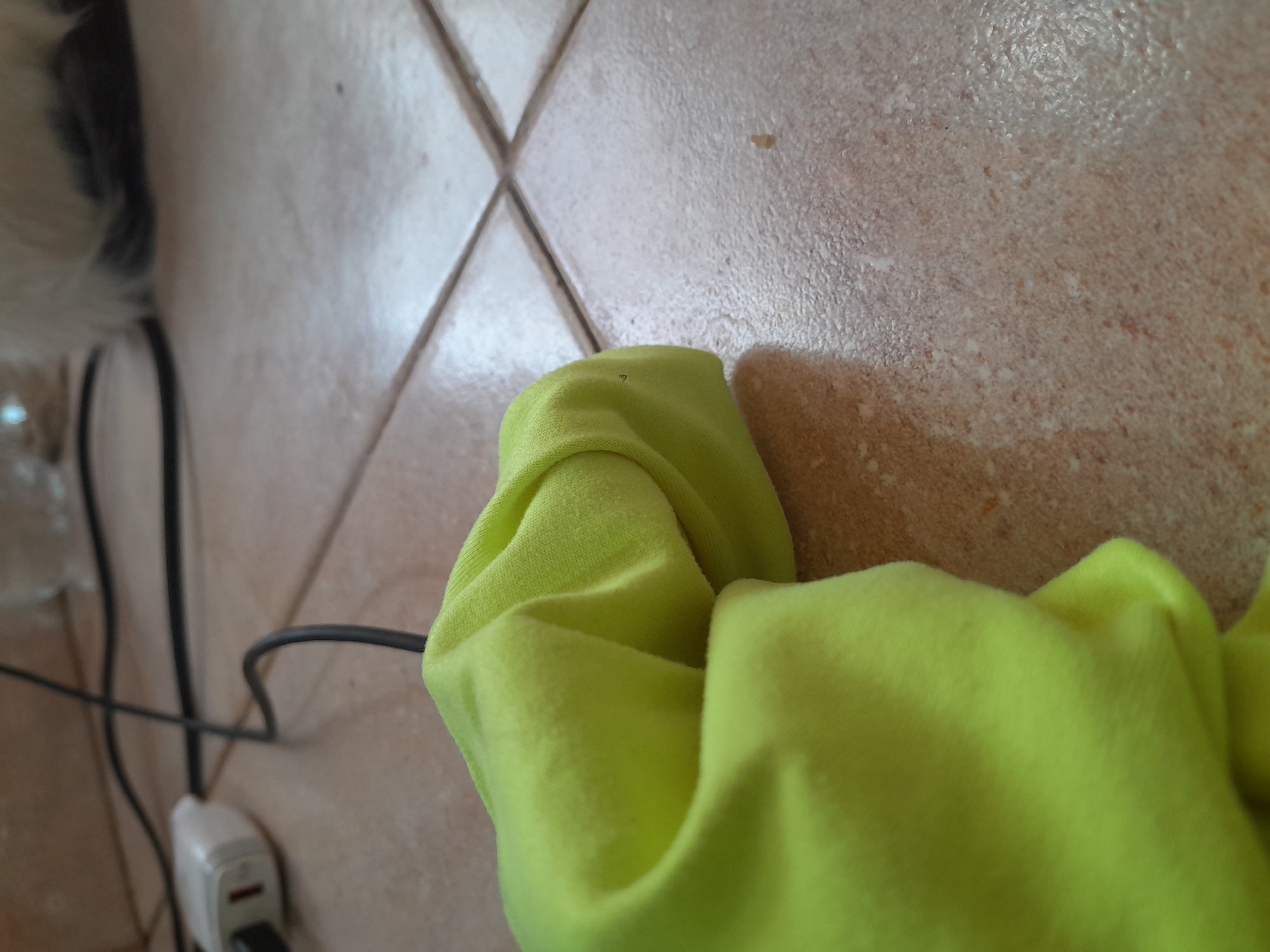 The image shows a close-up of a bright green piece of fabric, possibly a t-shirt or sweatshirt, bunched up on a tiled floor. The tiles are a mottled brown and beige color with visible grout lines. In the background, out of focus, is a white electrical plug connected to a black cord that runs across the floor. Further in the background, a blurry white and black object, possibly an animal, is visible. The scene appears to be indoors, likely in a residential setting in Santa Ana, Venezuela, given the tiled floor often found in homes in the region. There are no people visible in the image.