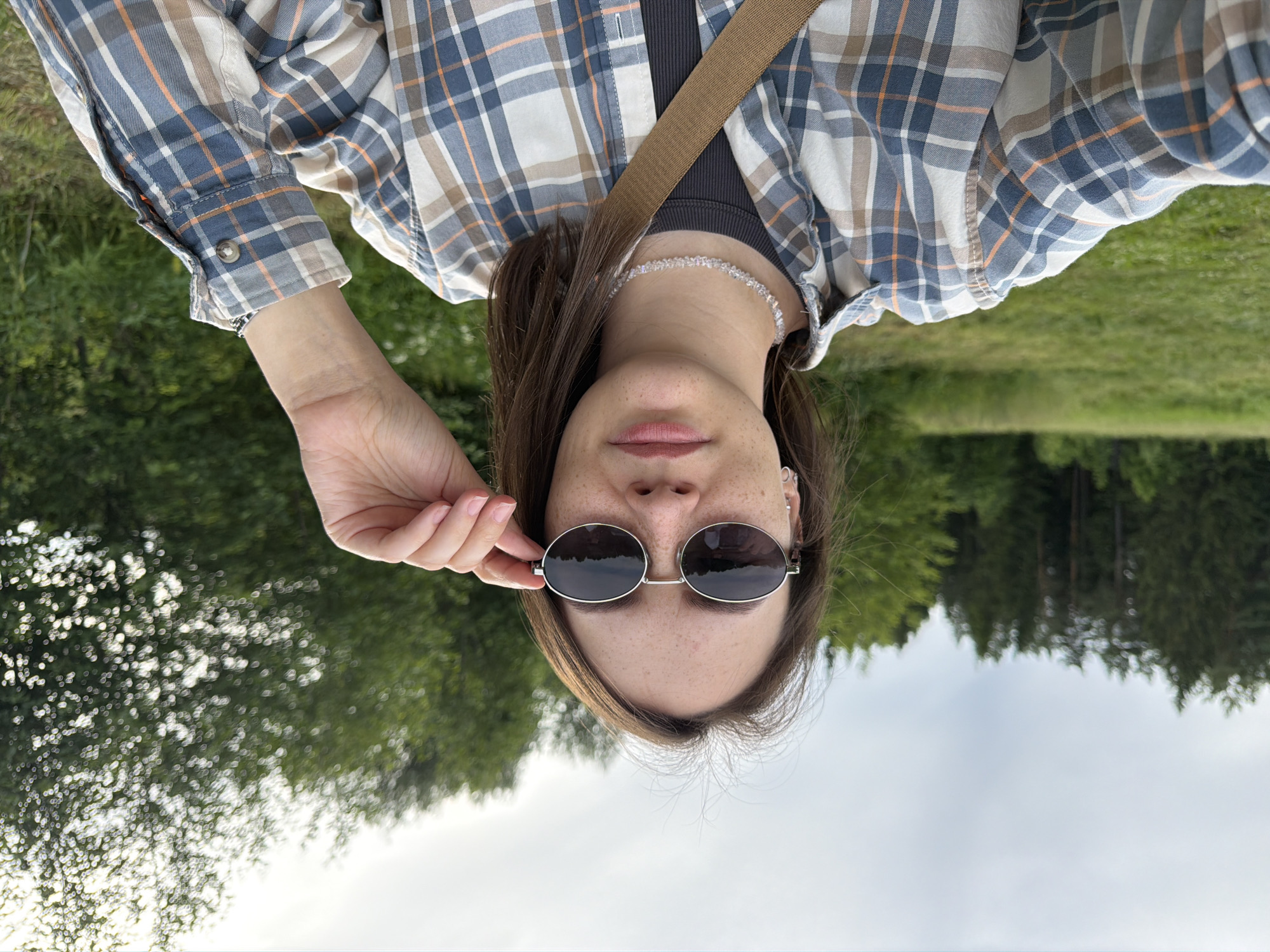 A woman is taking a selfie outdoors in Garevaya, Russia. She is wearing sunglasses, a plaid shirt, and a necklace. Her hair is brown and pulled back. The background is a field with trees. The sky is cloudy.