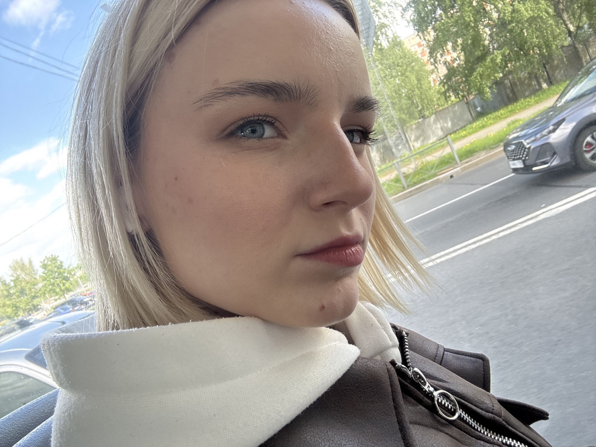A close-up, medium shot captures a fair-skinned woman with blonde hair, partially visible blue eyes, and natural lip color. She is looking to the right. She is wearing a white hoodie with a brown, faux leather jacket.
Outside the window is visible a light gray car driving along a street. In the background are green trees and buildings. The location is likely Saint Petersburg, Russia, based on architectural style and the license plate of the car.