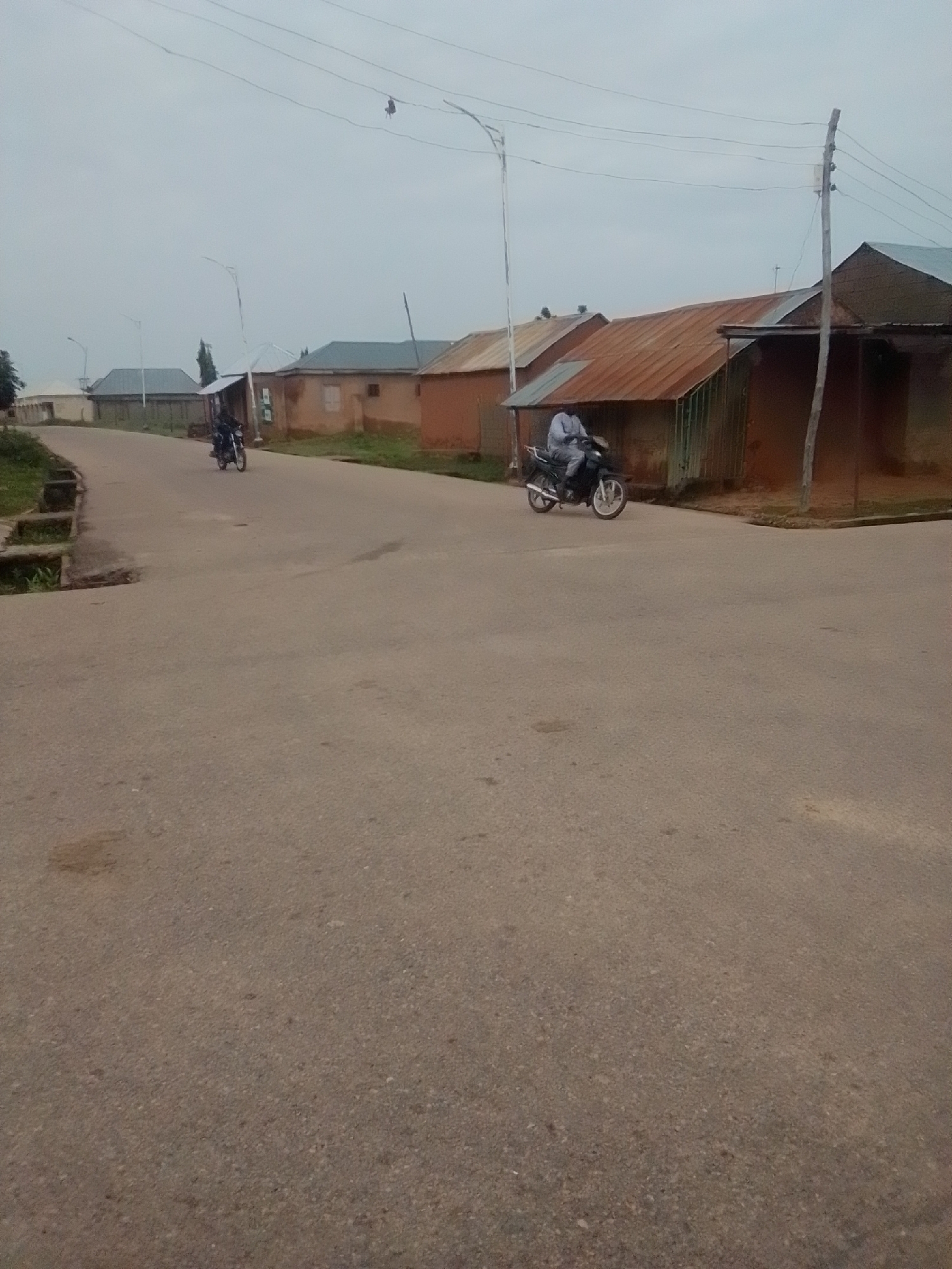 The image captures a street scene in Gombe, Nigeria, under an overcast sky. The foreground is dominated by a paved road with some discoloration and patches. On the left side of the road, there is a drainage channel with overgrown grass. In the middle ground, a person is riding a motorcycle on the road. Further back, a row of single-story buildings with corrugated metal roofs is visible. The buildings have reddish-brown walls and some appear to be under construction or in disrepair. Utility poles and wires are present, indicating the presence of electricity infrastructure. The overall environment suggests a somewhat unkempt but functional urban setting.
