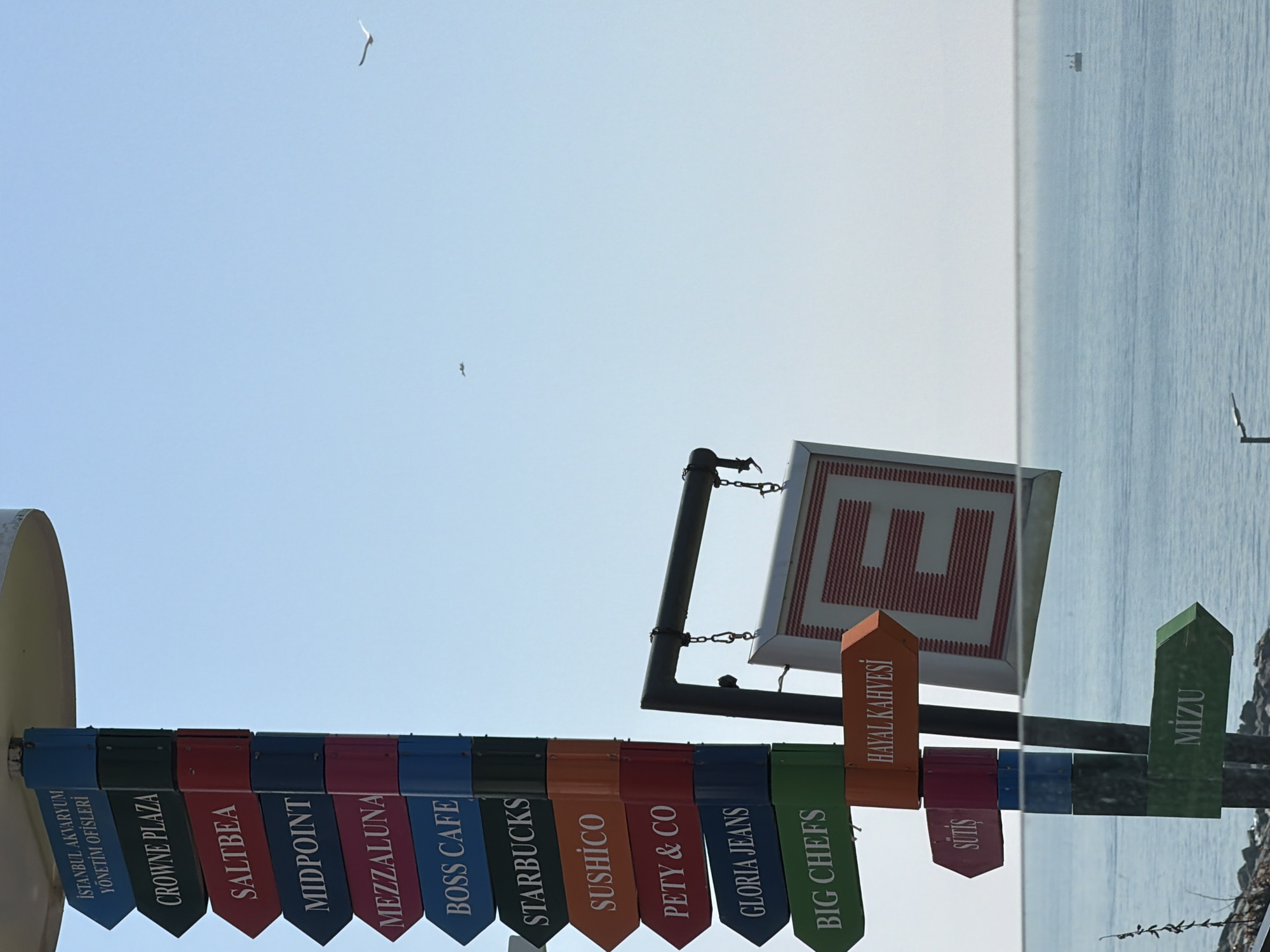 The image shows a directional sign in Bakırköy, Turkey, pointing to various commercial establishments like Istanbul Aquarium, Crowne Plaza, Saltbea, Midpoint, Mezzaluna, Boss Cafe, Starbucks, Sushico, Pety & Co, Gloria Jeans, Big Chefs, Sütis, Hayal Kahvesi, and MIZU. The sign features a white square with a red "E" design. The background includes a light blue sky, a body of water, and a boat in the distance. Two birds are flying in the sky. The signs are multi-colored, each with a different name of a business. The location is suggested to be near the waterfront in Bakırköy.
