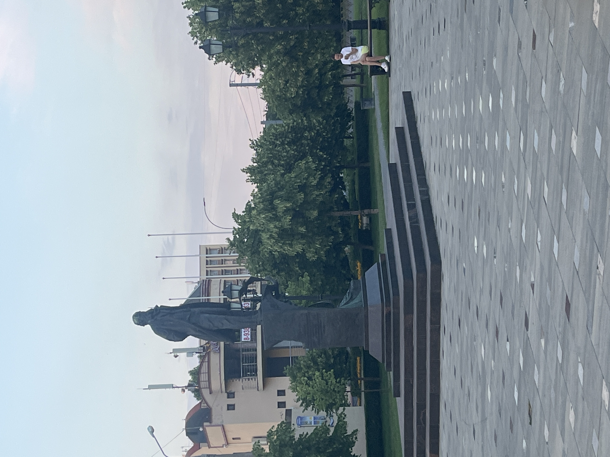 The image depicts a public square in Krasnodar, Russia, dominated by a large bronze statue of a man, likely a historical figure, on a granite pedestal. The statue stands on a tiered base with wide steps. In the background, several multi-story buildings are visible, some with balconies and signs. Trees line the square, and lampposts are scattered throughout. A person is seated on a bench on the right side of the image, looking at their phone. The ground is paved with large square tiles, some lighter and some darker, arranged in a patterned layout. The sky is overcast with a light haze.