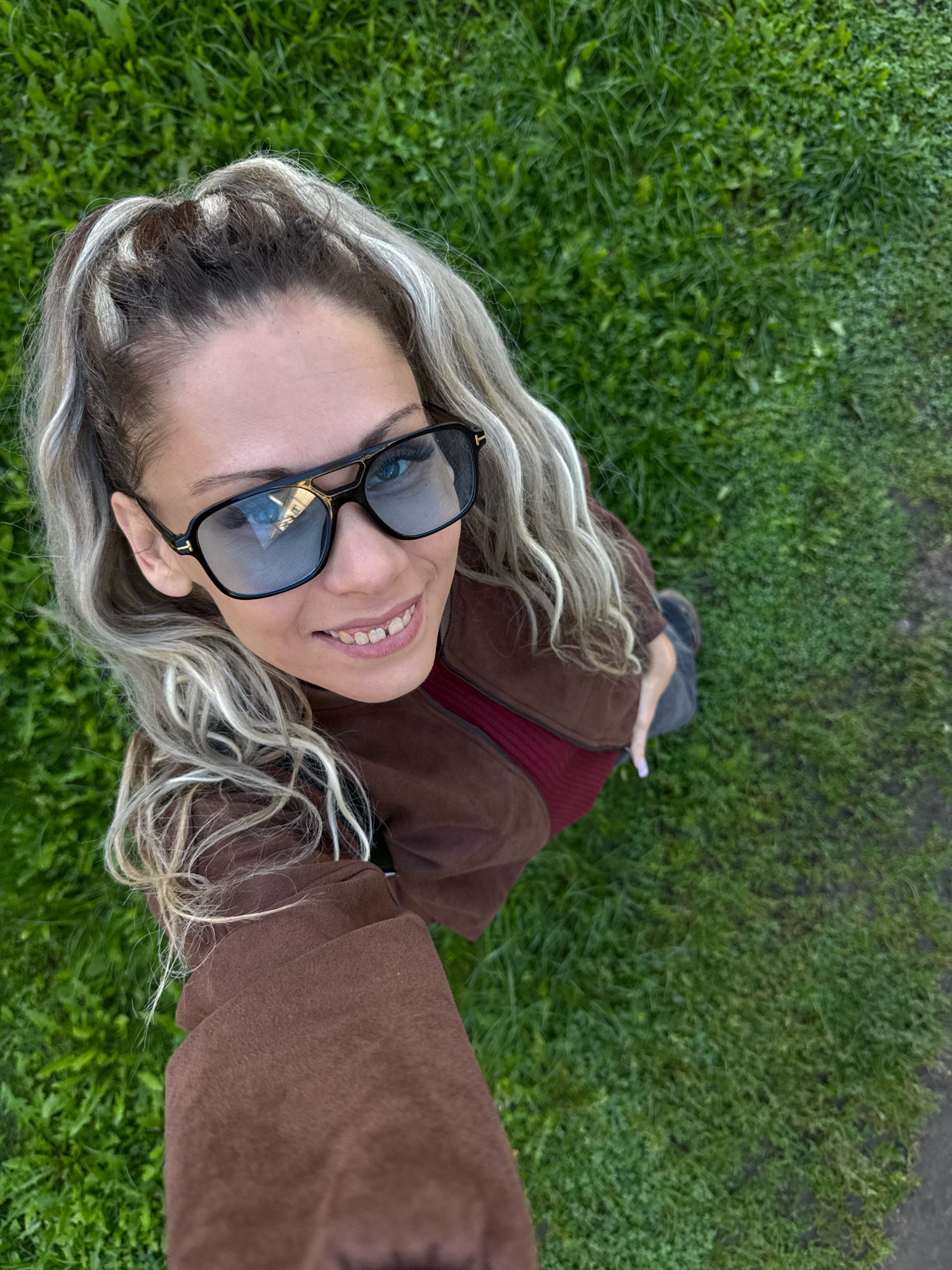 A high-angle, full-body selfie captures a woman with blonde, wavy hair pulled into a high ponytail and wearing large black sunglasses. She is standing on a grassy area with a dirt path visible to the right. The woman is wearing a brown jacket over a dark red top. Her right hand is extended, holding what appears to be a smartphone. She is smiling at the camera. The background is lush green grass. The image is taken outdoors, likely in a park or a residential area, possibly in the city of Rusanovo, Russia.