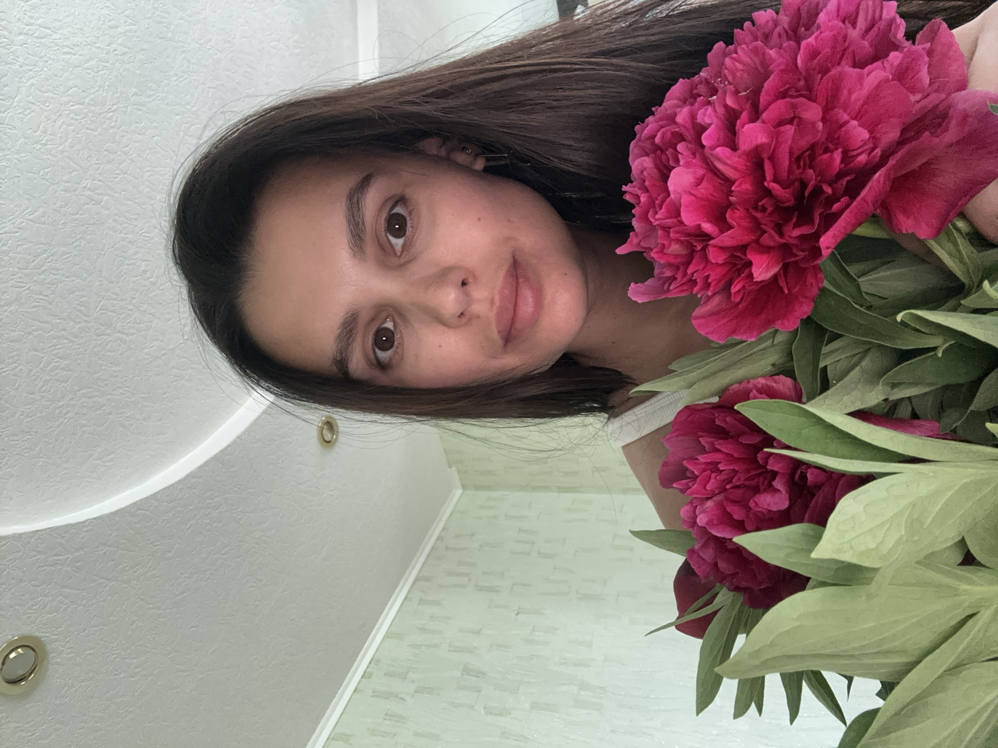 A young woman with long brown hair is holding a bouquet of bright pink peonies. The background appears to be an interior setting with a white ceiling and patterned light-green walls. There are two light fixtures visible in the ceiling. While a location in Chaikovsky, Russia is mentioned, there is no visual information in the image to confirm this detail.