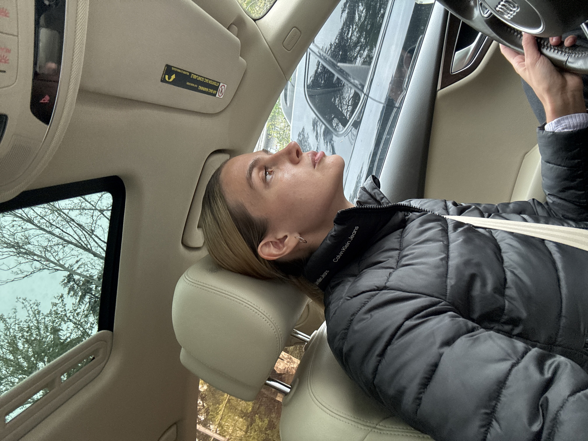 The image captures a candid moment inside a car, likely an Audi given the steering wheel logo, with a young woman in the driver's seat. She is seen from the right side, reclined back with her head resting against the light-colored headrest, looking upwards with a neutral, contemplative expression. Her blonde hair is visible, and she wears a small hoop earring. She is dressed in a black puffer jacket, with "Calvin Klein Jeans" visible on the collar, and a striped shirt cuff peeking from beneath her jacket sleeve. Her right hand is lightly gripping the steering wheel.
The car's interior is predominantly light beige or cream, including the headliner, sun visor, and dashboard elements. Through the side window and sunroof, bare tree branches and sparse green foliage against an overcast sky are visible, suggesting an outdoor, possibly park-like, setting in Tbilisi, Georgia, during a season like late autumn, winter, or early spring.
Notable textual details include "AIR BAG WARNING" and "ATTENTION SAC GONFLABLE" (with a safety symbol) on the sun visor, and "AIRBAG" on the A-pillar. The steering wheel prominently displays the Audi rings logo. The overall lighting suggests it is daytime under an overcast sky. The woman's relaxed posture indicates she is either resting, waiting, or not actively driving at the moment.