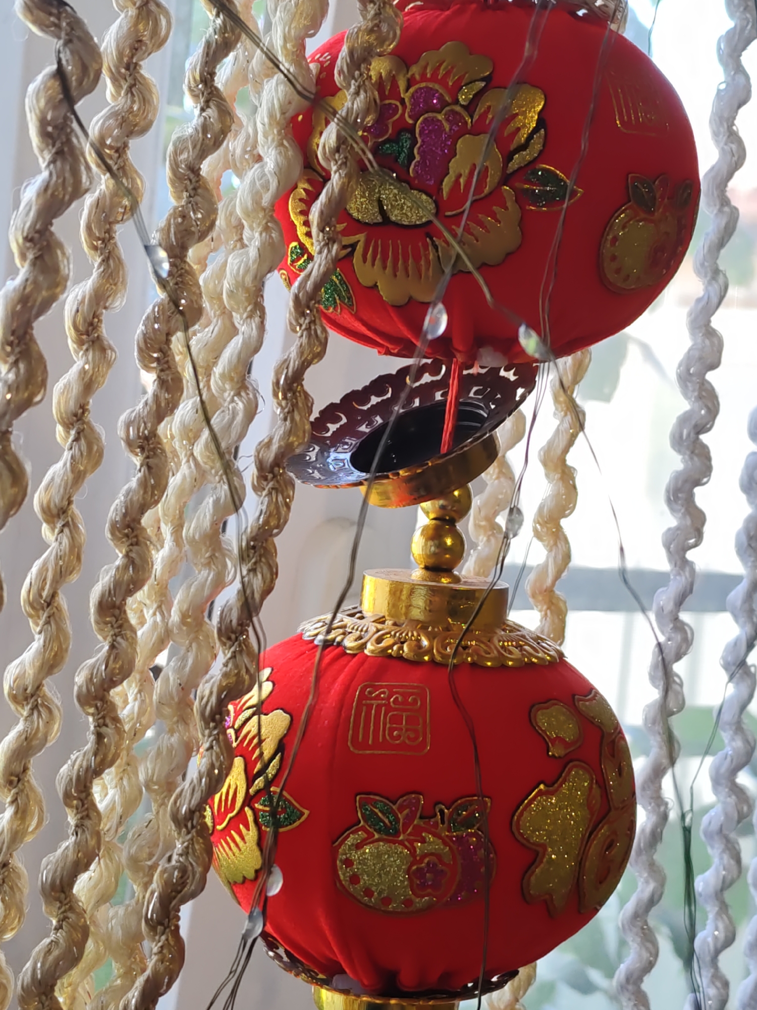 Two red Chinese lanterns are suspended by decorative ropes. The upper lantern has a floral design with gold and pink accents, while the lower lantern features gold lettering and a fruit motif. Both lanterns are adorned with small, reflective lights. The background consists of a blurred window with sunlight filtering through, and hanging white decorative elements. The image does not contain any recognizable landmarks or features that would indicate its location in Rostov-on-Don, Russia.