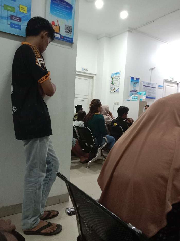 The image depicts an indoor waiting area within a public facility in Gladaksari, Indonesia. The room features white walls, a light-colored tiled floor, and overhead lighting. On the left, a young adult male stands, leaning against a white wall, facing away from the camera. He wears a black short-sleeved shirt with yellow detailing on the sleeves, light blue jeans, and dark flip-flops. His arms are crossed at his chest.
In the midground to the right, a row of individuals is seated on black and chrome waiting chairs. One person is visible from the back, wearing a dark green top and blue jeans. Another person behind them wears a light-colored head covering (hijab). Further right, another individual wears a black shirt with yellow accents. A light brown or beige head covering (hijab) belonging to a person in the immediate foreground partially obscures the view of the seated individuals.
The walls are adorned with various informational posters; one on the far left is primarily blue and white with a QR code, while another poster further right has a green and white design. Two white doors are visible in the background, and a light blue counter or cabinet, labeled "KOTAK SARAN" (suggestion box), is present in the far background on the right.