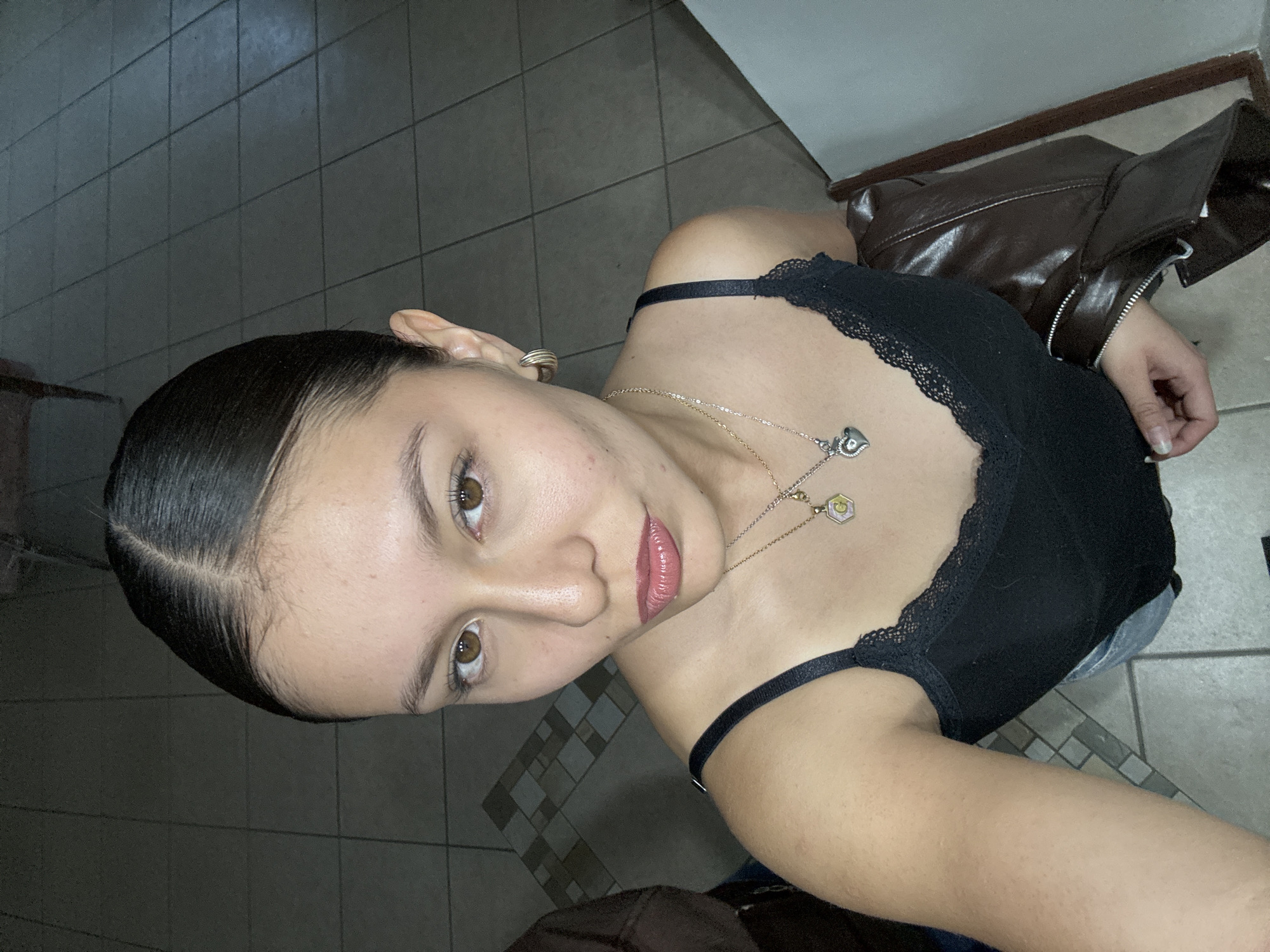 A woman with dark hair pulled back stands in what appears to be a tiled room. She is wearing a black top with lace trim and has a gold-colored necklace with pendants. Behind her is a brown jacket. The location is suggested to be Heroica Puebla de Zaragoza, Mexico.