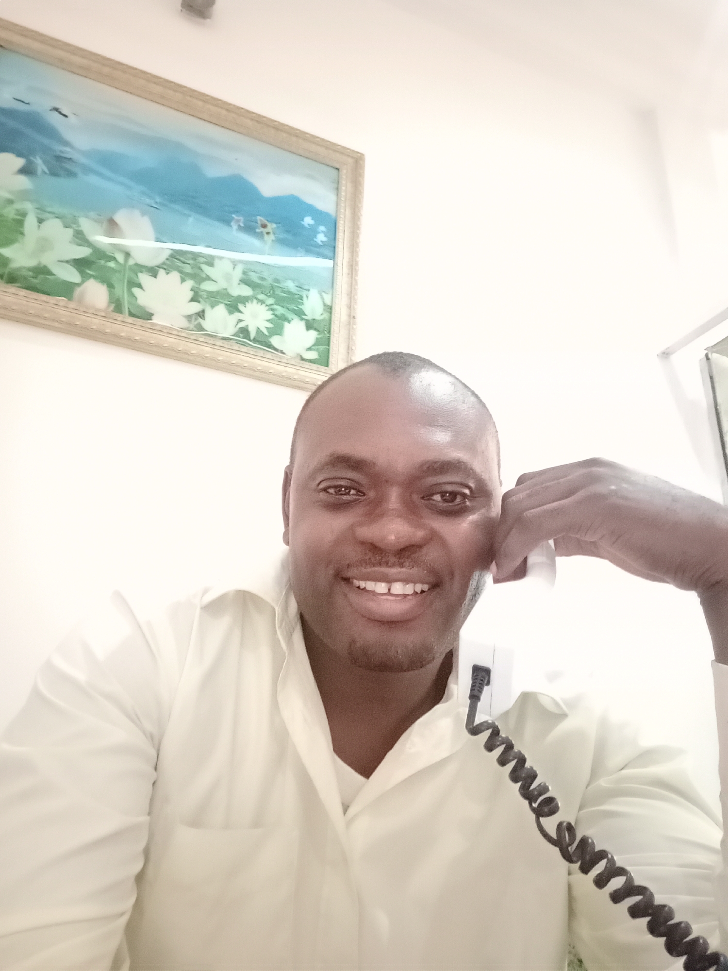 A man is on the phone in an indoor setting. He is smiling and wearing a light-colored, button-down shirt. A landline phone is positioned near his ear, the coiled cord visible. Behind him is a framed picture of a floral scene with mountains in the distance. The location is not specified as Gwarinpa, Nigeria, but implied by the user’s request.