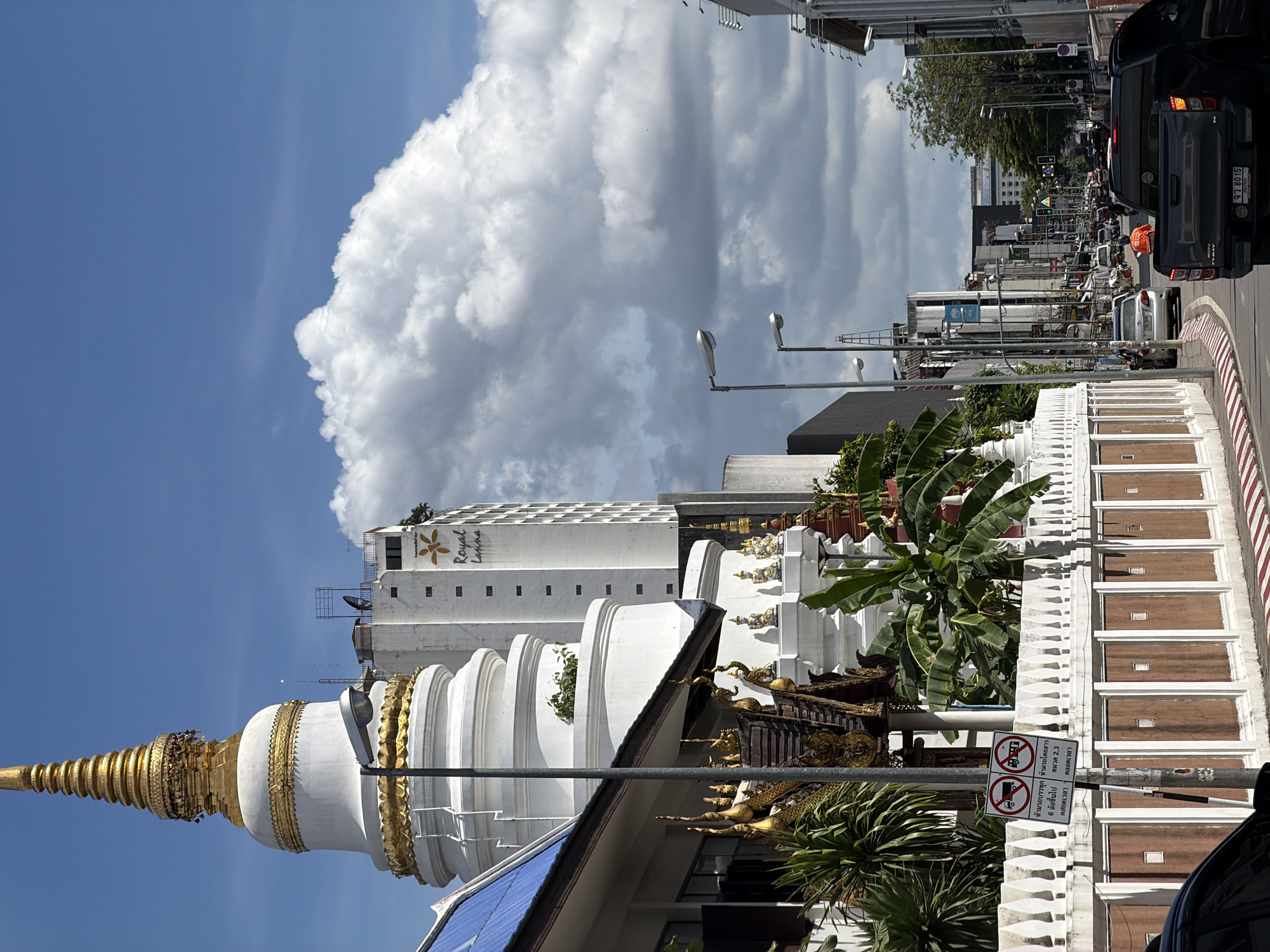 This is a vertical, eye-level, medium shot of a street scene in Chiang Mai, Thailand, dominated by a large, white, multi-tiered stupa with gold accents in the foreground. The stupa's tiered structure is characteristic of Lanna architecture, and it is adorned with intricate golden detailing. Behind the stupa, a tall, modern white building with a floral logo that reads "Royal Lanna" is visible. The sky is a clear blue, with a large, fluffy white cloud taking up a significant portion of the upper left side of the frame.
The street in the background is busy with traffic, including cars and motorcycles. Traffic lights and street lamps line the road. Signs with Thai text are visible, indicating regulations and restrictions. One sign shows symbols for prohibited trucks and buses, with text "ห้ามรถบรรทุก" (No trucks), "ห้ามรถโดยสาร" (No buses), "6 ล้อขึ้นไป" (6 wheels and up), and "ตลอดเวลา" (All the time) in Thai. Another sign also indicates "ตลอดเวลา" (All the time) along with "หมวด 2,3" (Category 2,3). A license plate on a dark-colored SUV reads "จว 8015" (JW 8015).
The scene suggests a bright, sunny day, likely in the morning or afternoon, given the lighting. The presence of lush green foliage, including banana trees, adds to the tropical feel of the setting. The overall atmosphere appears to be a blend of historical architecture and modern urban life, typical of Chiang Mai.