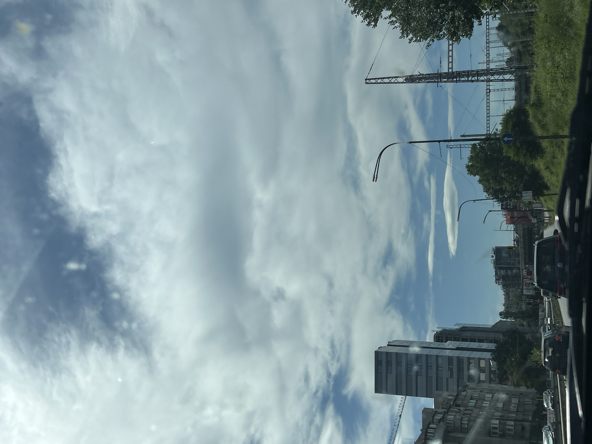 The image, taken from within a vehicle, shows a street scene in Sofia, Bulgaria. Several modern high-rise buildings, constructed primarily of concrete and glass, are visible to the right. Streetlights are present along the roadway, and a railway infrastructure including overhead power lines and support structures is observed in the background. A traffic arrow sign is also visible on one of the traffic signs. Traffic consists of parked cars. The sky is partly cloudy with a blue sky background. The image is oriented vertically, suggesting it was taken with a mobile device.