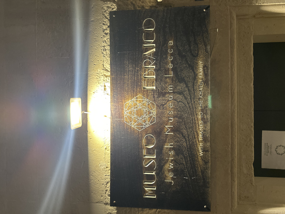 The image displays the illuminated entrance sign of the Jewish Museum Lecce (Museo Ebraico Lecce) at night, located in Lecce, Italy. A rectangular, dark-colored sign with a wood grain texture is centrally positioned on a light-colored, rough-hewn stone wall. The words "MUSEO EBRAICO" in large, gold-toned, raised letters are prominently displayed at the top, with "Jewish Museum Lecce" in smaller, similar lettering below. A complex geometric design, possibly a stylized Star of David, is situated between "MUSEO" and "EBRAICO". Below the museum name, "WITH THANKS TO THE KHALILI FAMILY" is visible in smaller text.
A single, rectangular, wall-mounted floodlight is positioned directly above the sign, casting a bright, warm yellow light onto it and the wall below. Distinct light streaks and subtle rainbow-colored lens flares emanate from the light source, extending upwards and to the sides. The sign is secured to the stone wall by four visible screws or bolts, one in each corner. Below the sign, the top portion of a dark-colored doorway is visible, framed by the same light-colored stone. A white paper notice, partially visible on the door, indicates "INAUGURAZIONE 24 GENNAIO 2023 ORE 18:00". No individuals are present in the scene.