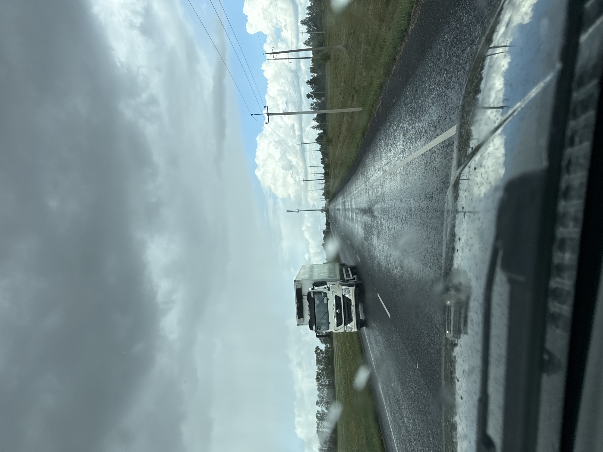 A white refrigerated truck is driving on a wet asphalt road. The sky is overcast with dark clouds. The road is flanked by green fields and scattered trees. There are utility poles along the side of the road. The image appears to be taken from inside a vehicle, with raindrops visible on the windshield. The scene is set in a rural area, possibly in or near Novyi Buzets, Russia.