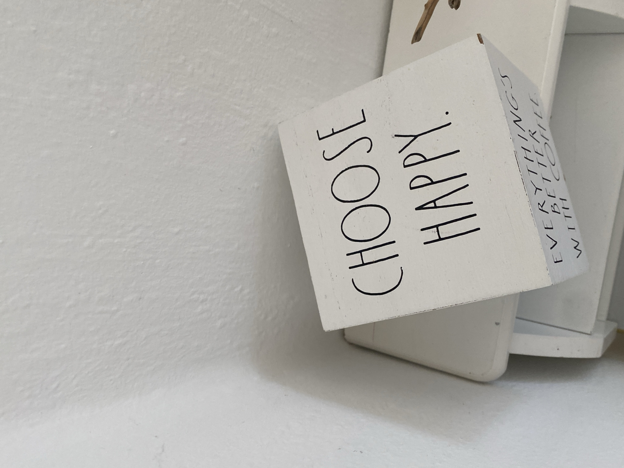 A white wooden sign with black lettering sits on a white shelf against a white textured wall. The sign has two lines of text on its visible face: "CHOOSE HAPPY." On the angled side of the sign, the text reads "EVERYTHING'S BETTER WITH COFFEE." A small, light-brown twig rests to the right of the sign. The scene is indoors, with soft lighting that casts a shadow on the wall to the left. The location is described as the city of Bad Sachsa, Germany.
