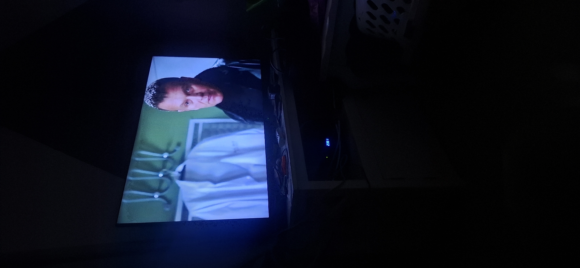 The image shows a television screen displaying a man with short, spiky hair and a stern expression. He is wearing a dark, high-collared garment, possibly a coat or jacket, with a fur or textured trim. Behind him, on the screen, is a white lab coat hanging on a rack against a green background. The television is placed on a white table or stand, which also holds some electronic devices and cables. A digital display on one of the devices shows "2331". The overall scene is dimly lit, suggesting it is nighttime or the room is dark. There is no discernible connection to Yerocham, Israel in the visual content of the image.