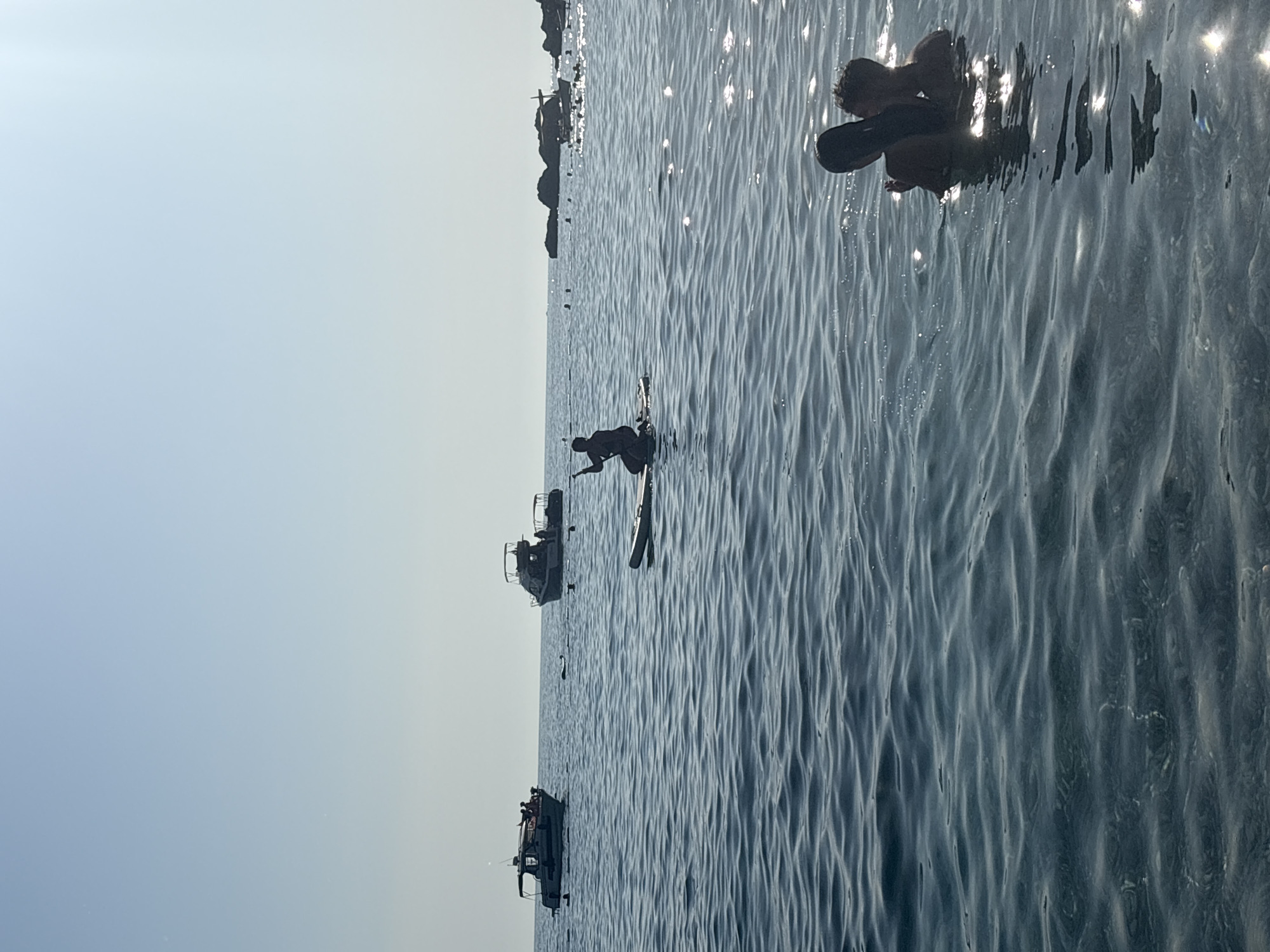 In the city of Drobnići, Montenegro, on a clear day, two people are submerged in the calm, shimmering sea. The water is a deep blue, with light glinting off its surface. In the foreground, to the right, two individuals are partially visible, their heads and shoulders breaking the water. One has long dark hair, and the other is a child or smaller person. They appear to be interacting, possibly embracing or playing.
Further out in the sea, a person is silhouetted on a paddleboard, kneeling with a paddle in hand, seemingly preparing to move across the water. In the middle distance, two boats are anchored or cruising. The boat on the left is smaller, with several people visible on its deck. The boat on the right is larger, also with passengers. Scattered across the water are numerous buoys or markers. In the background, rocky formations rise from the sea. The sky is a pale, clear blue, with no clouds visible. The overall impression is a peaceful scene of recreation on a sunny day.