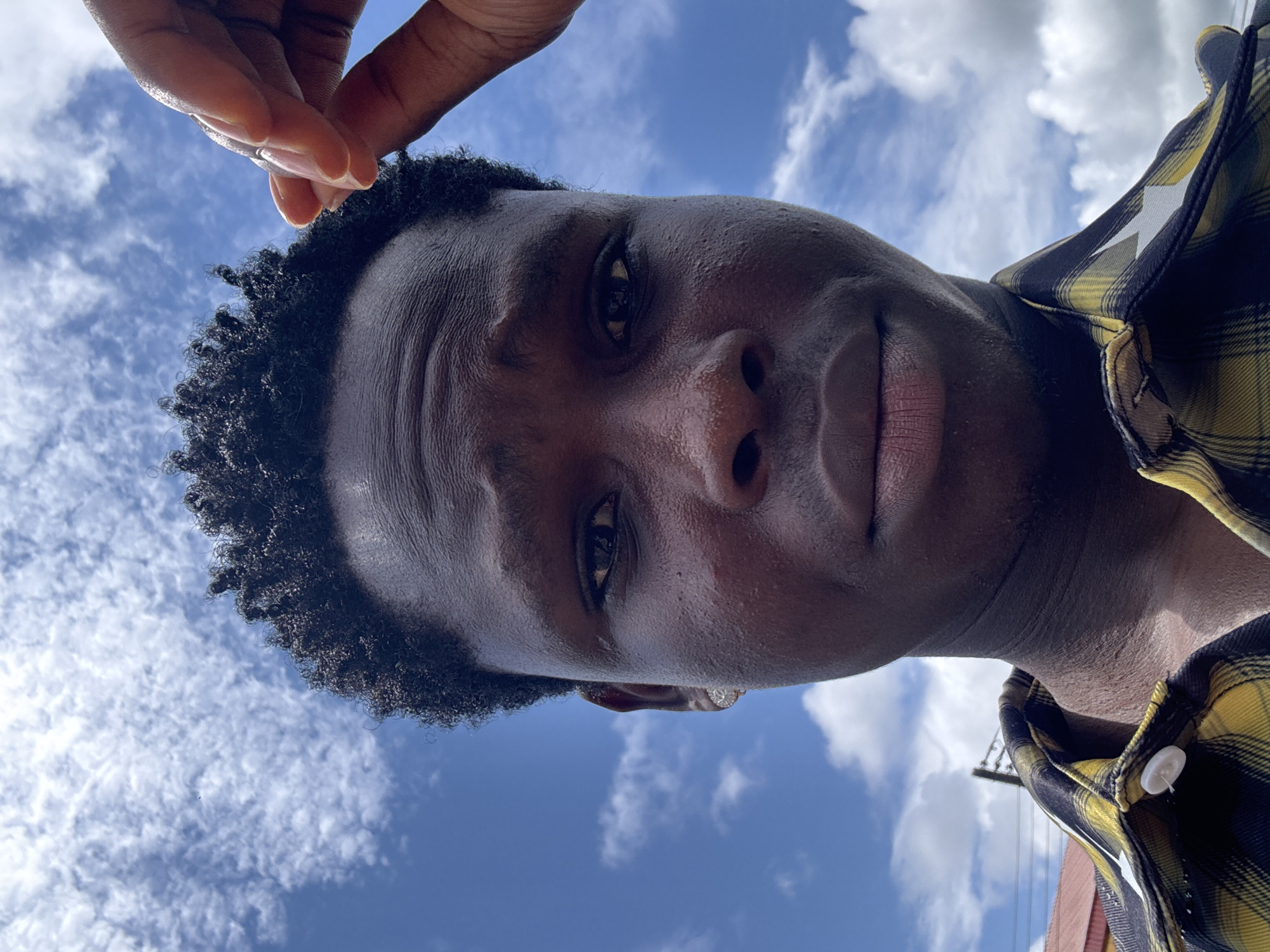 A medium shot, taken outdoors on a bright day, shows a close-up of a young Black male. He is facing the camera, looking forward with a neutral expression. His dark, curly hair is cut short on the sides and slightly longer on top. His skin is dark and appears slightly dewy. He is wearing a yellow and black plaid shirt with white buttons. His right hand is raised to his forehead, with his index finger touching his hair. The background is a clear blue sky dotted with white, fluffy clouds. In the lower left of the frame, a portion of a red-roofed building and a utility pole are visible, suggesting an urban or semi-urban setting. The image is a selfie, taken from a low angle. There is no indication of the specific location within Ihiagwa, Nigeria.