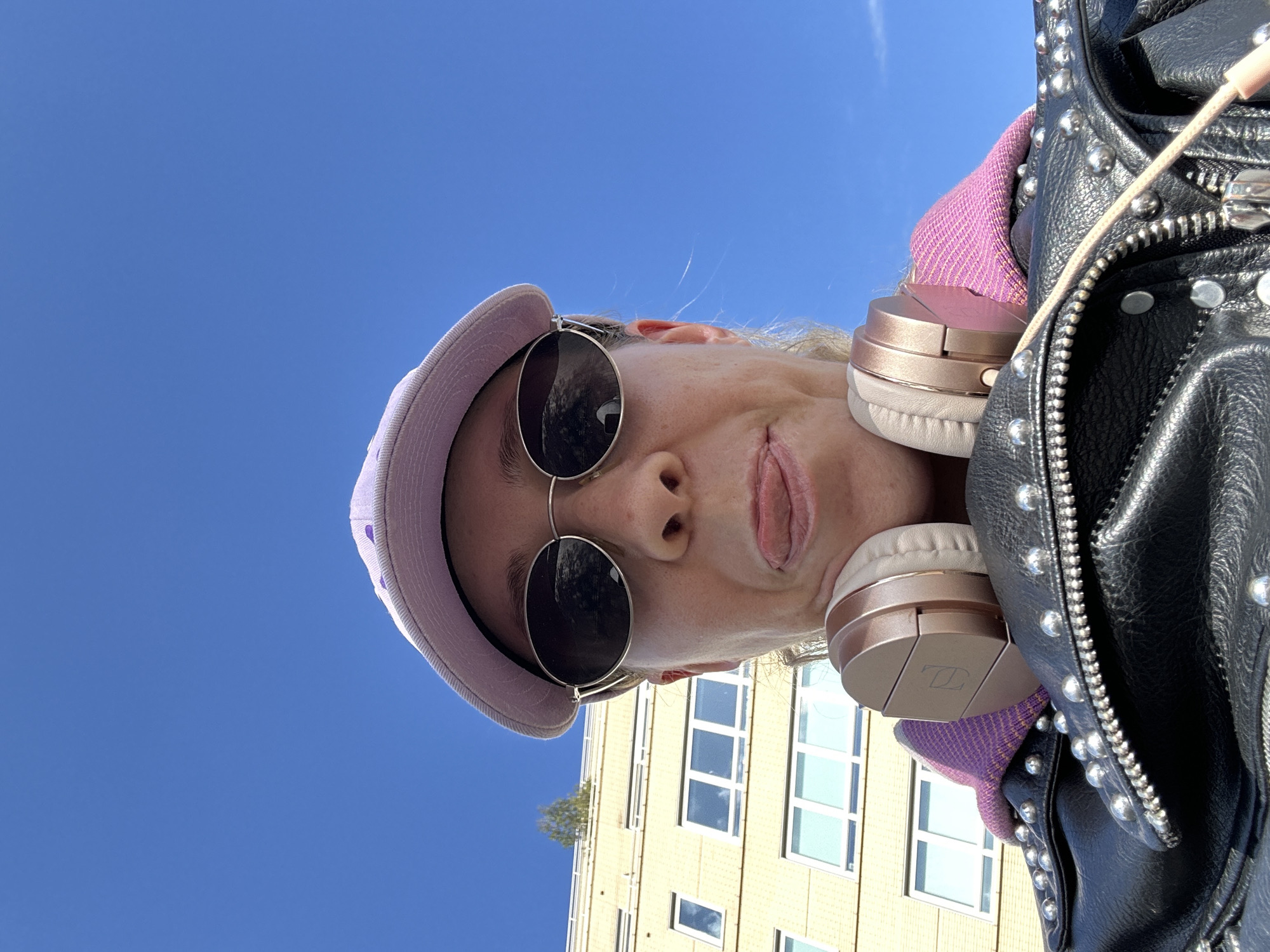 A selfie portrait, shot from a low angle, features a woman wearing a light purple cap and sunglasses, with rose-gold headphones around her neck and a black studded leather jacket. She is looking down at the camera, with her tongue slightly sticking out. Behind the woman, the blue sky is visible. Additionally, a multi-story building with multiple rows of windows, light yellow in color, is present in the background. While the photo provides visual context of an outdoor setting, there is insufficient information in this image to conclude that the location is specifically Amsterdam, Netherlands.