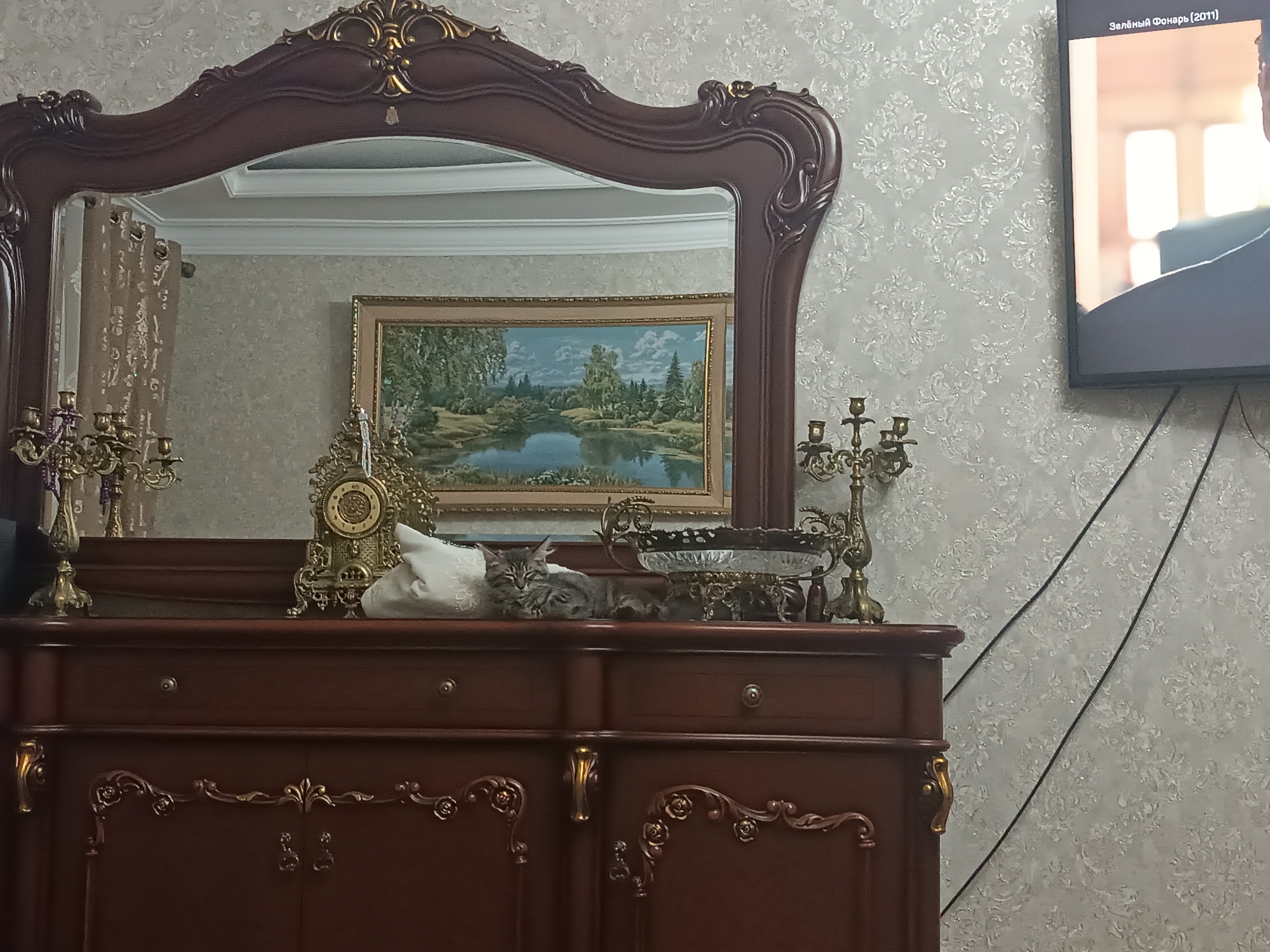 The media file shows a domestic scene in Izberbash, Russia. A vintage wooden dresser with ornate gold detailing serves as the focal point, topped with various decorative objects. A large, ornate mirror hangs above the dresser, reflecting a framed landscape painting of a serene lake surrounded by trees. On the dresser, a golden clock with intricate carvings sits next to a white fabric object, possibly a cushion or folded cloth. To the right of these items, a small grey tabby cat is resting, with its head tilted and eyes partially closed. Further to the right, on the dresser's surface, is a decorative glass bowl with a metal stand, and a brass candelabra. To the far left of the dresser, another brass candelabra is visible. The wall behind the dresser is covered in textured wallpaper with a floral pattern. To the right of the dresser, a flat-screen television is mounted on the wall, displaying a still image of a movie scene with Russian text at the top: "Зелёный фонарь (2011)". The lighting suggests it could be daytime, possibly indoors with artificial lighting. There are no visible weather cues or strong emotional indicators beyond the relaxed posture of the cat.