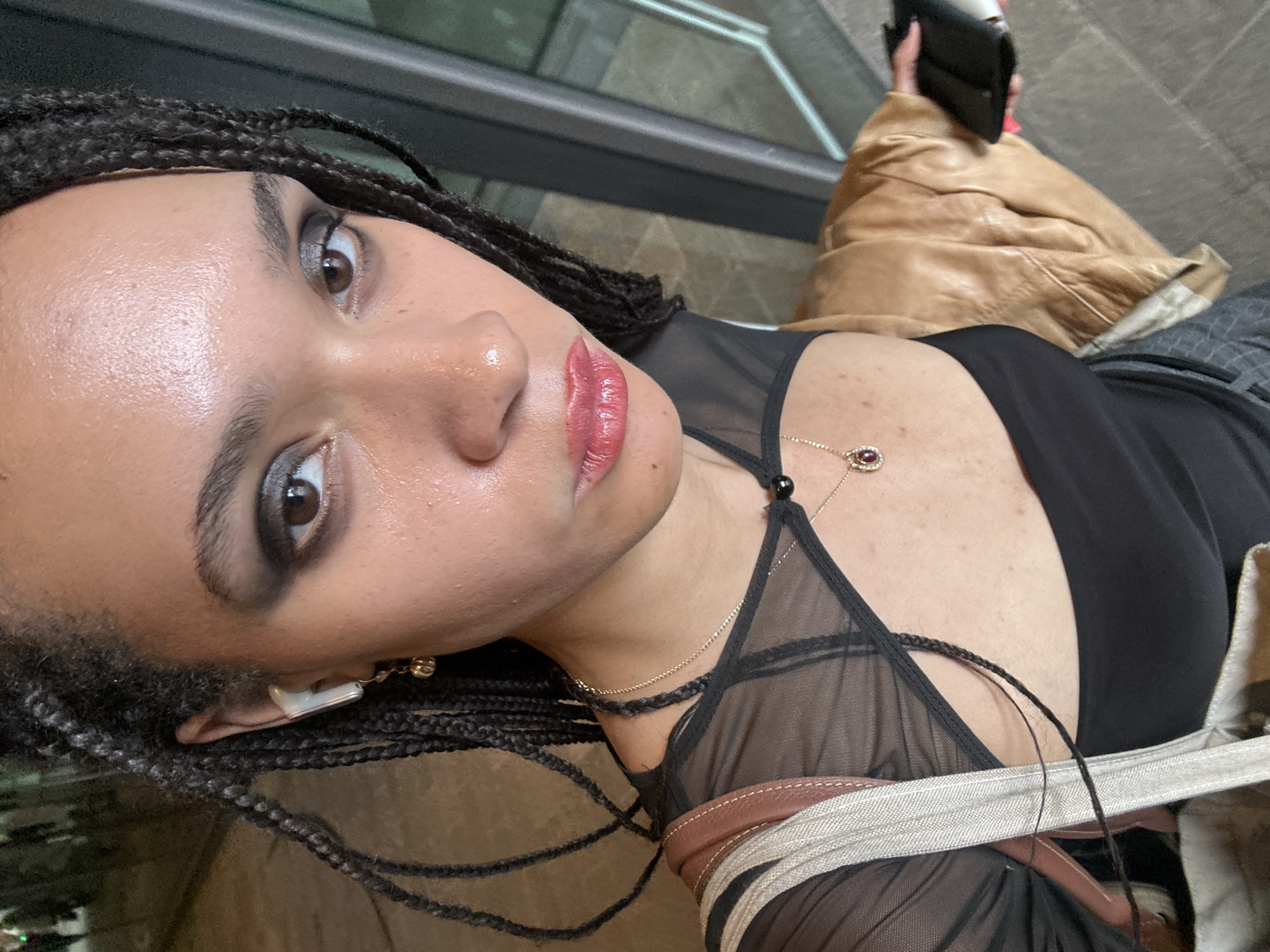 A woman with dark skin and braided hair is facing the camera. She is wearing a black top with a sheer neckline and has on makeup, including dark eyeshadow and lipstick. She has jewelry including a gold necklace and earrings. A tan leather jacket is partly visible in the background. She is outside, and a building is visible. This appears to be in London, United Kingdom.