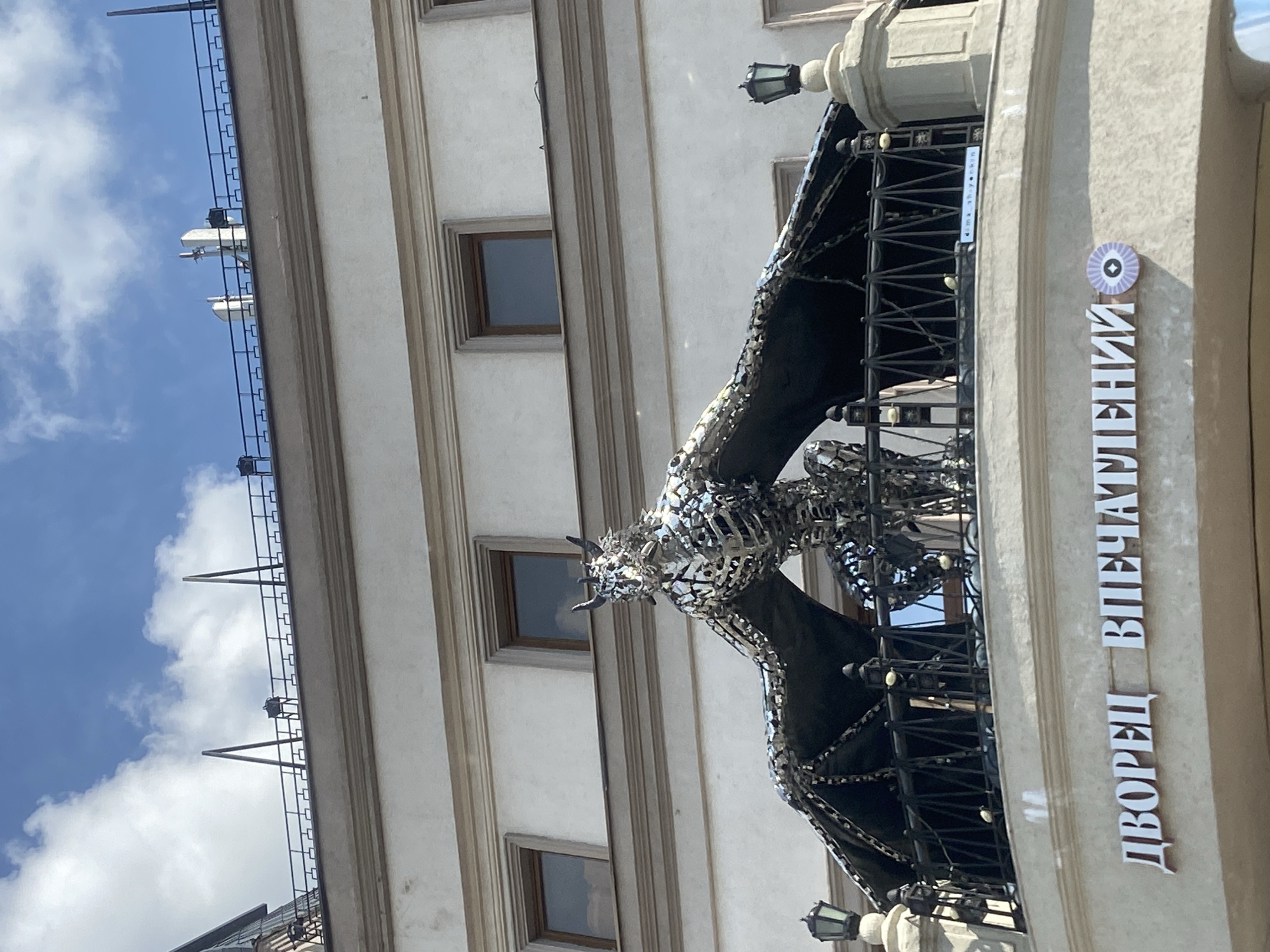 The image shows a metal sculpture of a dragon with outstretched wings, perched on a balcony overlooking a building with the Cyrillic inscription "ДВОРЕЦ ВПЕЧАТЛЕНИЙ" (Palace of Impressions). The sculpture is made of reflective metal pieces and appears to be a decorative element. The building itself is light-colored with several windows and ornate architectural details. The sky above is blue with scattered white clouds. The sign indicates the location is in Kazan, Russia, as "Дворец Впечатлений" is a known attraction in Kazan. There are no people visible in the image. The scene is captured from a low angle, looking up at the sculpture and the building.