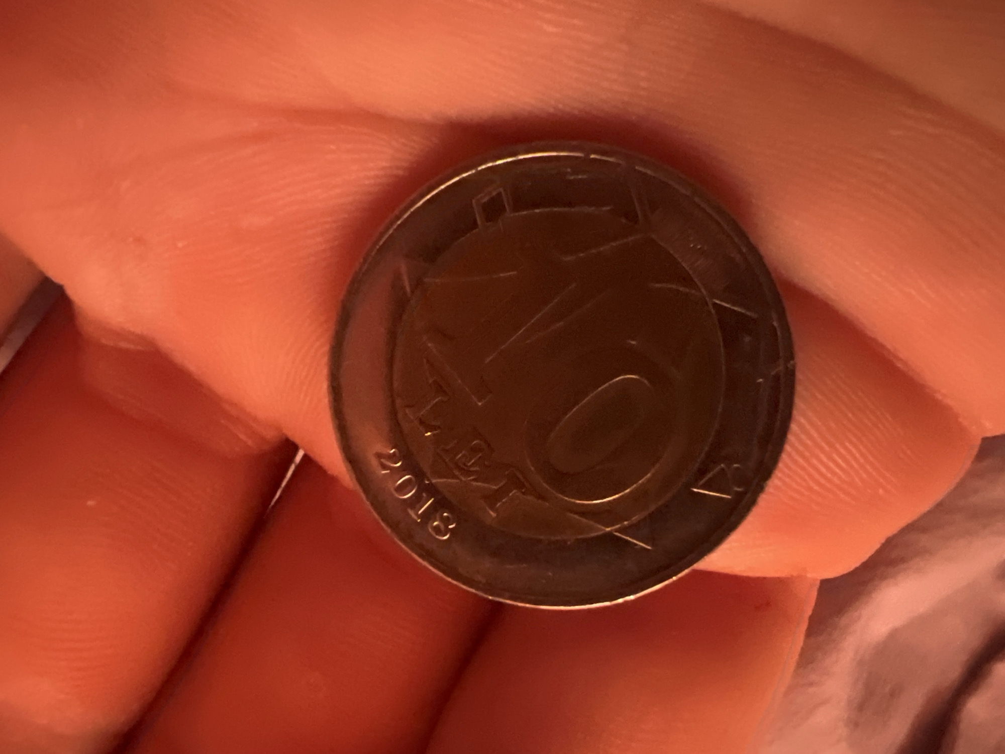 The image shows a close-up of a hand holding a coin. The coin appears to be a 10 cent Euro coin from Latvia, as indicated by the inscription "10 EIROPAS CENTI" and the Latvian coat of arms. The year "2018" is also visible on the coin. The hand is a human hand, with visible skin texture and creases. The background is out of focus but appears to be a warm, reddish-orange color, possibly fabric or skin. The lighting is warm, creating a glow around the coin and hand. There is no information in the image to suggest a specific location in the city of Ciorescu, Moldova.