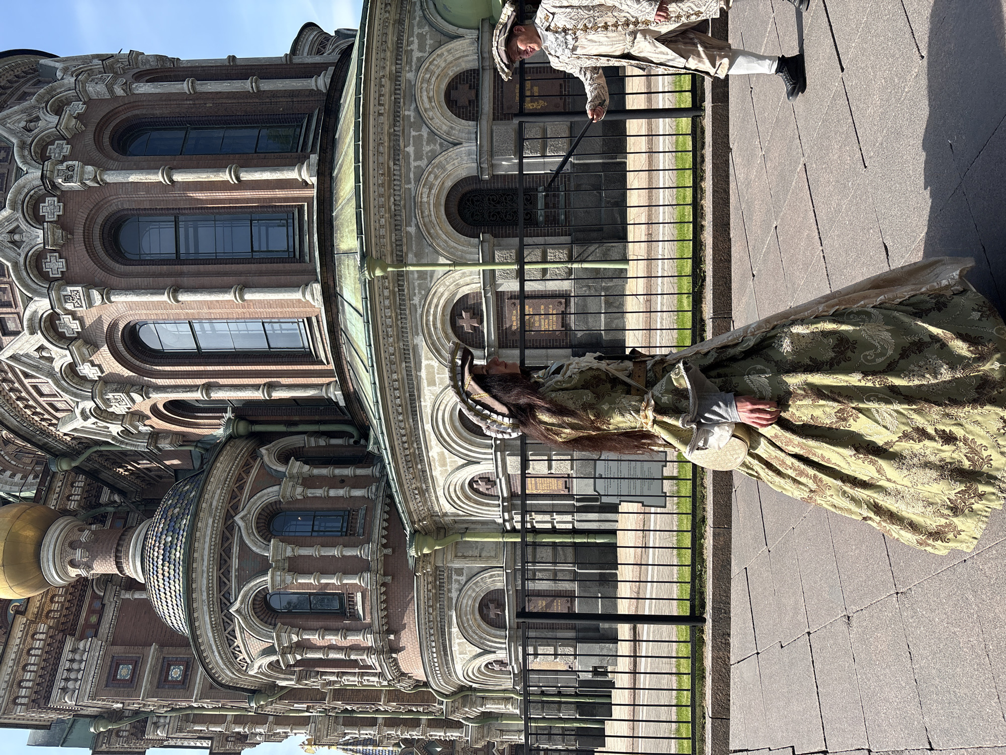 The image depicts two individuals dressed in historical attire in front of the Church of the Savior on Spilled Blood, located in Saint Petersburg, Russia.
A woman in a light green and gold patterned gown and ornate headwear stands facing the camera. A man in a white and gold patterned suit and hat is partially visible on the right.
The background features the elaborate architectural details of the Church, including its domes, arched windows, and decorative elements. A black metal fence is present in the foreground, separating the subjects from the church building. The ground is paved with large rectangular stones. The sky is visible in the upper left corner, suggesting a sunny day.