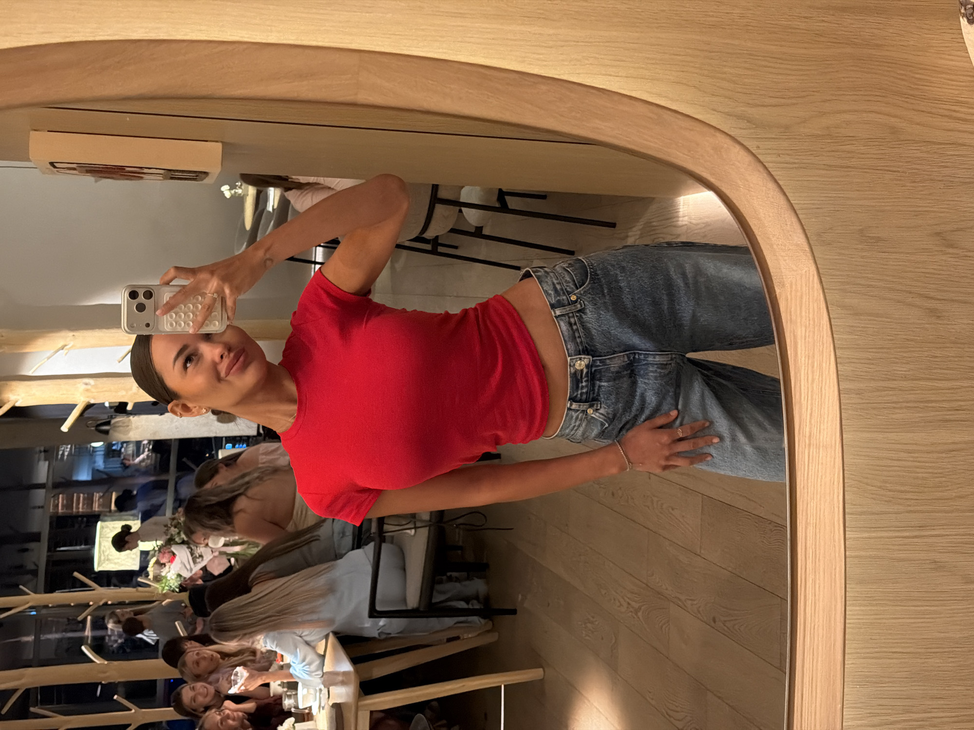 A woman takes a mirror selfie in a restaurant, she is wearing a red crop top and blue jeans. In the background, other people are seated at tables. The restaurant appears to have a modern, minimalist design with light wood accents. There is no information to determine the location in the city of Krasnoyarsk, Russia.