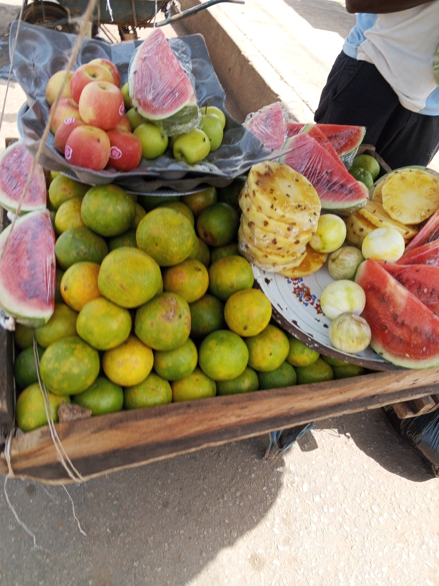 The image shows a street vendor in Bukuru, Nigeria, selling a variety of fruits from a wooden cart. The cart is filled with oranges, apples, watermelons, and pineapples. Some of the fruits are sliced and wrapped in plastic. A person is visible in the background, wearing a white t-shirt with blue trim and black shorts. The scene appears to be outdoors, with a paved surface and shadows cast by the sun. The cart is positioned on the side of the road, suggesting a typical street market environment. The overall impression is one of a vibrant, bustling marketplace.