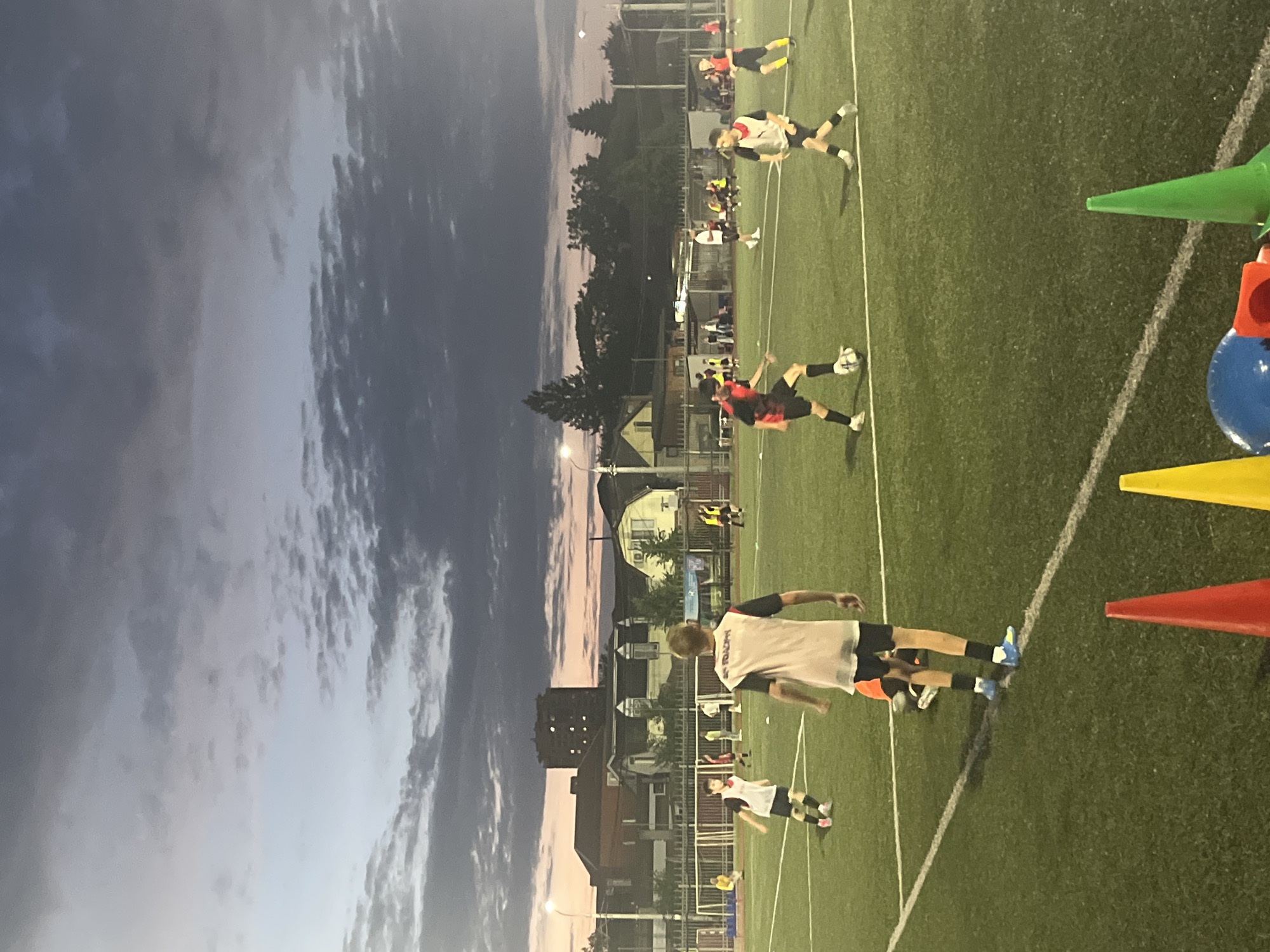 This is a vertical, low-angle outdoor photograph of a soccer game at dusk. The sky is a gradient of deep blue to soft pink and gray, with clouds scattered across it. The setting is a soccer field with green artificial turf and white boundary lines. Several young people, likely teenagers or young adults, are playing soccer. They are wearing athletic clothing, primarily black shorts and jerseys in various colors, including red, white, and yellow. Some players are in motion, kicking or running with the ball, while others are observing or standing on the field.
In the background, there are buildings and trees, suggesting an urban or suburban environment. The lighting appears to be a mix of natural twilight and artificial lights from the field. The overall mood seems to be one of active participation and a casual sporting event. There are several colorful cones (green, yellow, and red) and a blue object, possibly part of a training apparatus, visible on the right side of the frame. No visible text is discernible in the image.