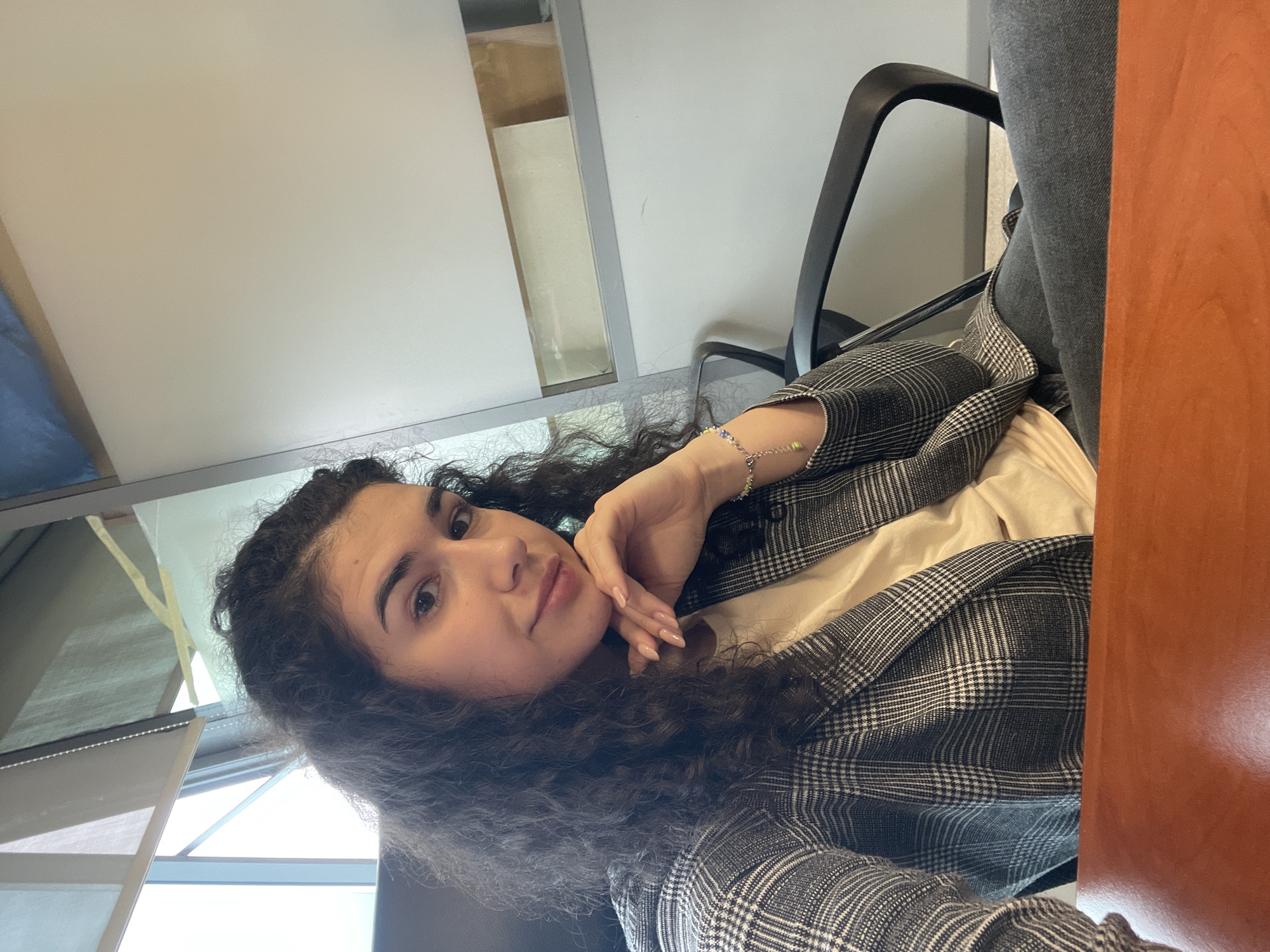 A young woman is captured in a selfie-style portrait, rotated 90 degrees counter-clockwise. She has dark, curly hair, medium skin tone, and is looking directly at the camera with a subtle, calm expression and a faint smile. Her right hand is gently placed near her chin. She is wearing a light-colored top under a grey and white plaid blazer, and grey pants or jeans are partially visible. A delicate bracelet with small, colorful beads adorns her right wrist.
The setting appears to be an indoor office or study environment, with an overhead white ceiling visible in the background. A black office chair is behind her, and to the right, a wooden or laminate desk edge is prominent in the foreground. Natural light streams in from a window or light source on the left side of the frame, suggesting it is daytime. The image contains no visible text. The location context provided is Sofia, Bulgaria.
