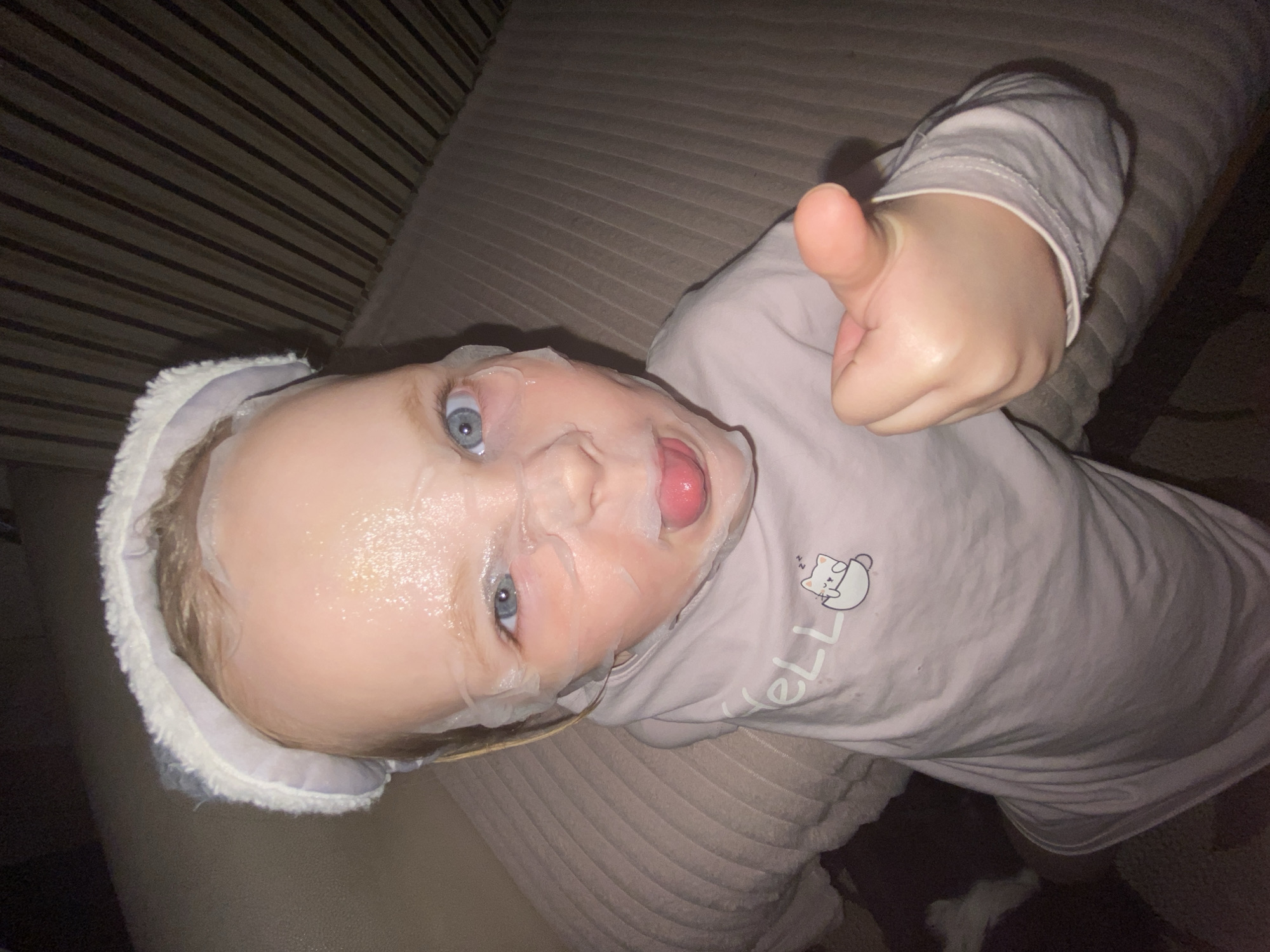 An overhead, slightly tilted shot captures a young child lying down, likely on a sofa or bed with a striped upholstered pattern. The child has fair skin and striking blue eyes, and their face is covered with a translucent white sheet mask, which has cutouts around the eyes and mouth. A fuzzy, light-colored headband is worn around their head.
The child's tongue is slightly out, adding a playful expression, and their left hand is raised, giving a clear "thumbs up" gesture. They are wearing a light pink or lavender long-sleeved shirt. On the shirt, a graphic depicts a white cat sleeping peacefully inside a teacup. Below the graphic, the partial text "ELL" is visible. The lighting is dim, suggesting an indoor setting, possibly in the evening. The scene conveys a humorous and endearing moment, likely of the child mimicking an adult's routine.