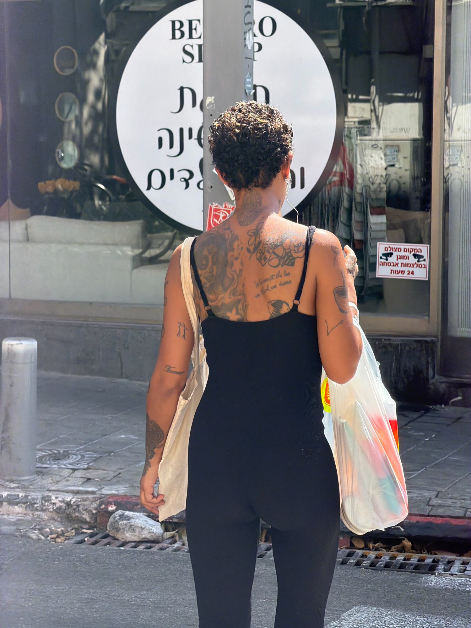 The image shows a woman with extensive tattoos on her back and arms, wearing a black sleeveless jumpsuit and carrying a canvas tote bag and a plastic shopping bag. She is walking on a sidewalk in Tel Aviv, Israel, with a storefront and a street visible in the background. The storefront has a circular sign with text in English and Hebrew, and a red and white sign with Hebrew text that appears to be a security notice. The woman is viewed from behind, and her face is not visible. The environment is urban, with a sidewalk, street, and building facades. The lighting suggests it is daytime.