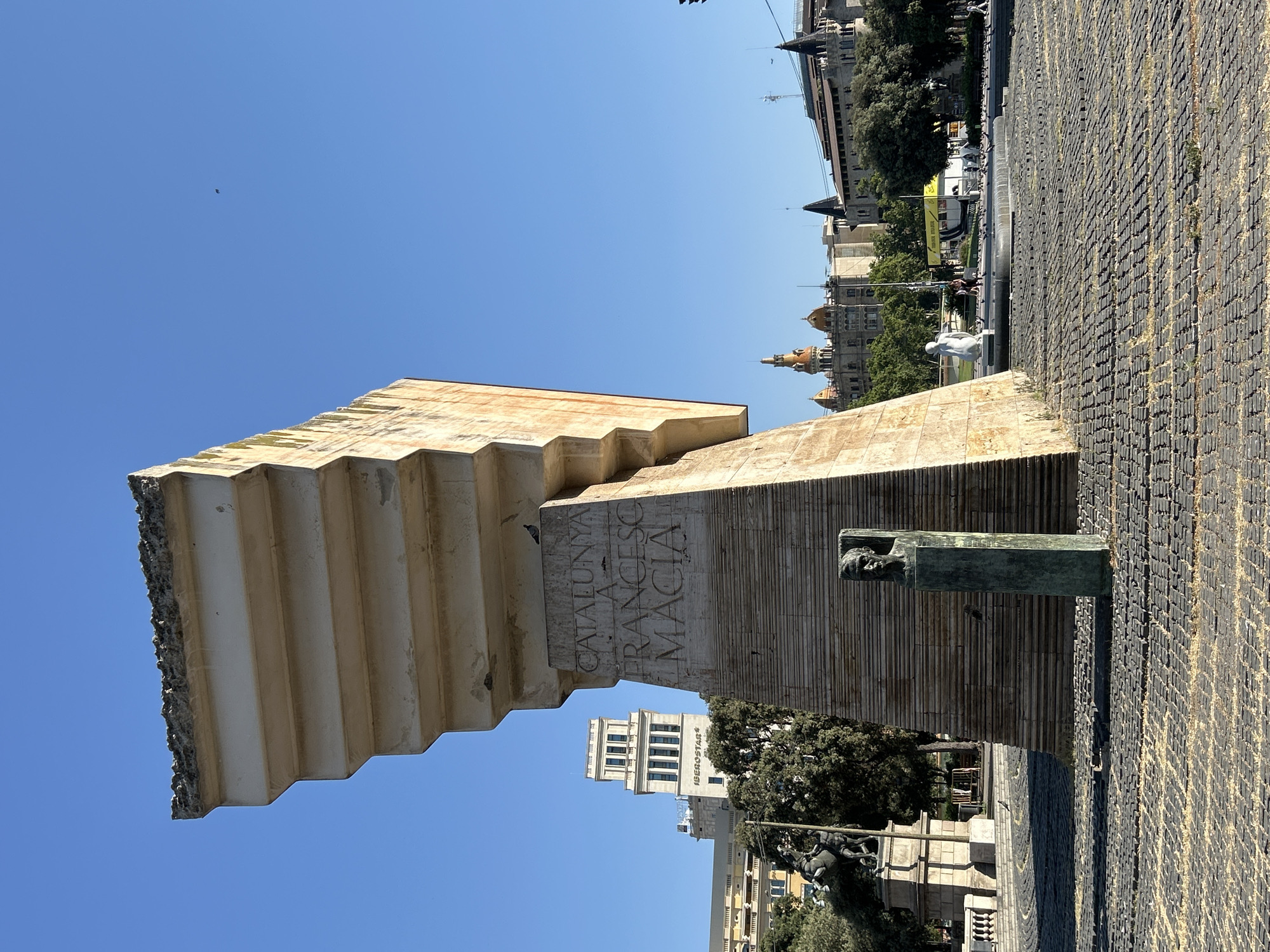The image captures a daytime view of the Monument to Francesc Macià in Barcelona, Spain, located in the Plaça de Catalunya. The monumental sculpture, constructed from stone, dominates the foreground. Its upper section is a massive, angular slab with a rough, eroded edge. The base of the sculpture is trapezoidal and inscribed with the words "CATALUNYA A FRANCESC MACIA". In front of the base is a bronze bust of a man, mounted on a rectangular pedestal.
The ground is paved with dark cobblestones arranged in a circular pattern, interspersed with patches of dry grass. To the left of the main monument, a statue of a man on horseback stands on a stone pedestal, partially obscured by trees. In the background, several buildings are visible, including a tall, white tower with the name "ISEBOSTAR" on it, and other structures with distinctive architectural features, such as domed roofs and spires.
The sky is a clear, bright blue with a few distant birds visible. In the middle ground, people are walking and a fountain is present, with several pigeons gathered around it. The overall scene is an outdoor public space bathed in sunlight.