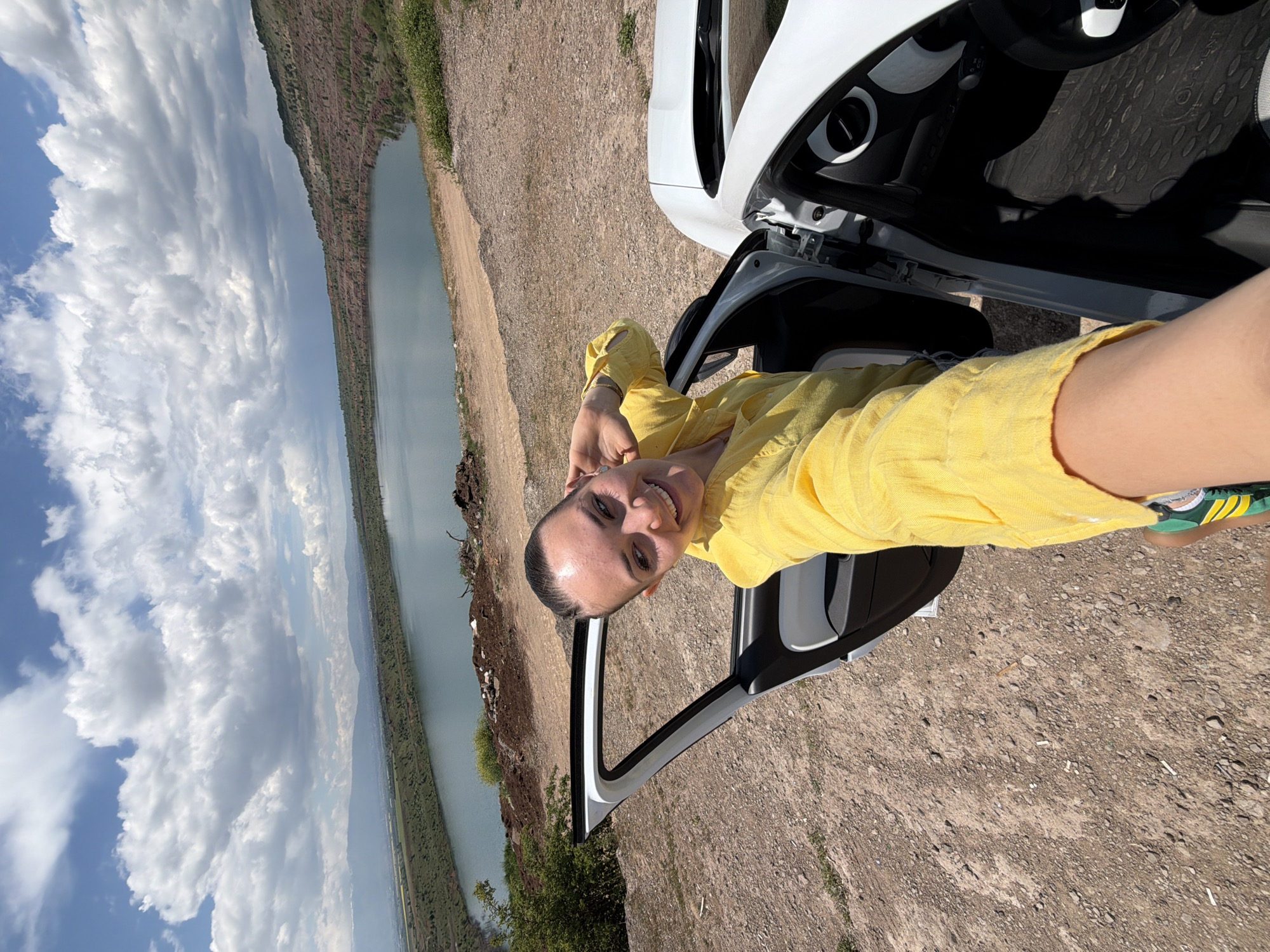 A person wearing a yellow shirt is standing by the open door of a white car in a rocky terrain. In the background, there's a body of water and a cloudy sky. The location appears to be an elevated area overlooking the water, possibly near a quarry or reservoir. There is no definitive information available in the provided image to identify the location as the city of Sofia, Bulgaria.