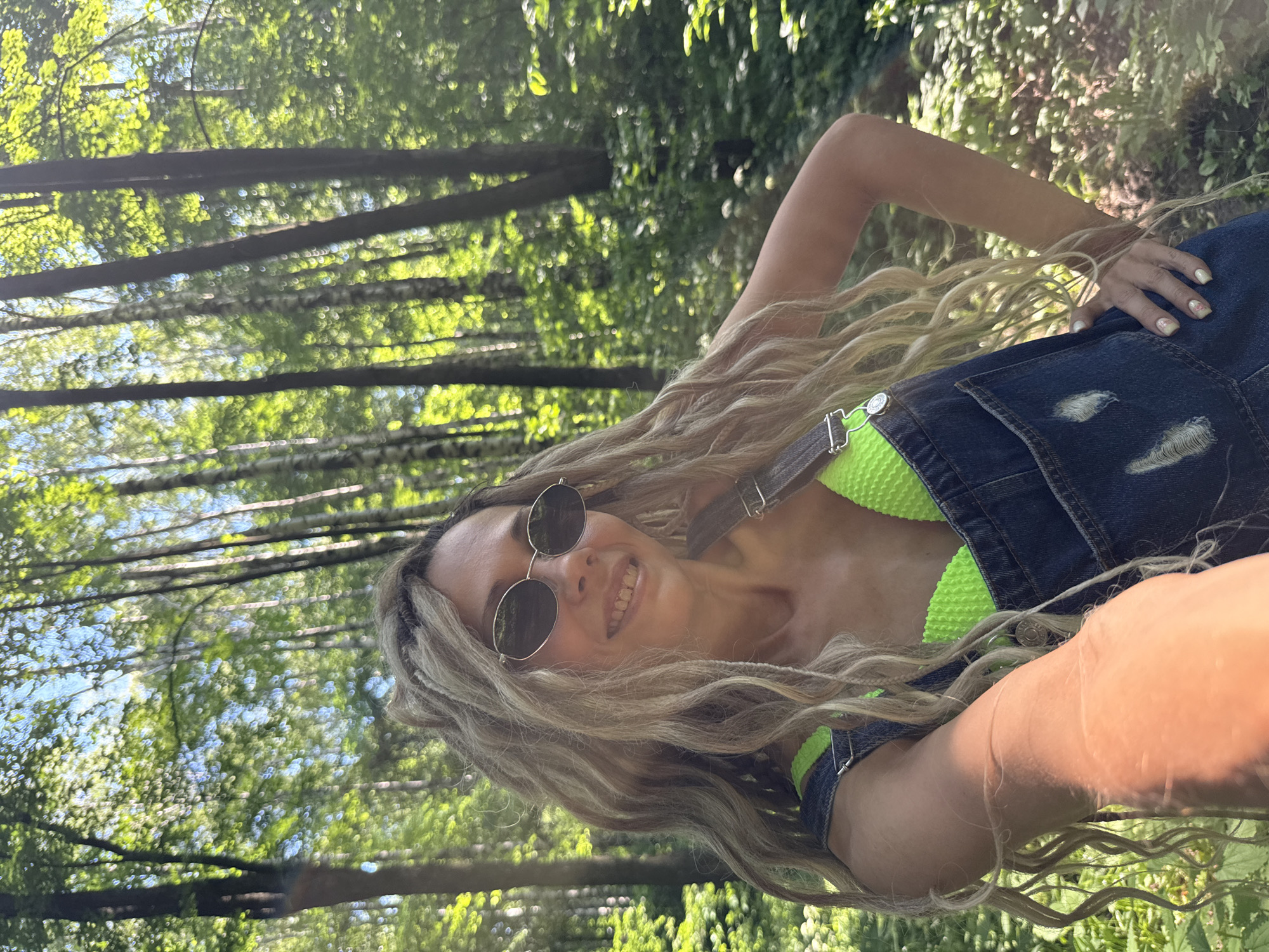 A woman with long, blonde dreadlocks and sunglasses smiles at the camera while standing in a forest. She is wearing a neon green bikini top and denim overalls. The sun is shining through the trees, creating a dappled light effect on the forest floor. The location is the city of Шебунино, Russia.