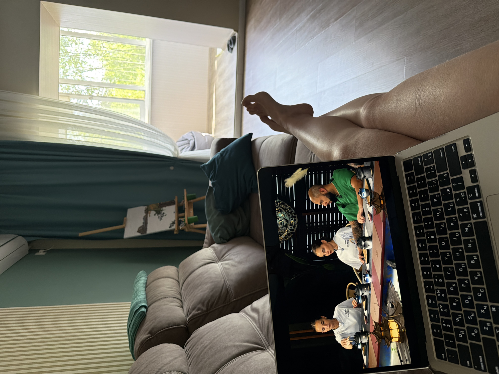 The image shows a person reclining on a sofa while watching a cooking show on a laptop. The laptop is positioned between the person's legs, with the keyboard visible. The cooking show features three individuals in a professional kitchen setting. In the background of the room, there's a window with a view of trees, and an easel with a canvas. The interior design includes neutral tones and a modern aesthetic. The location is not specified.