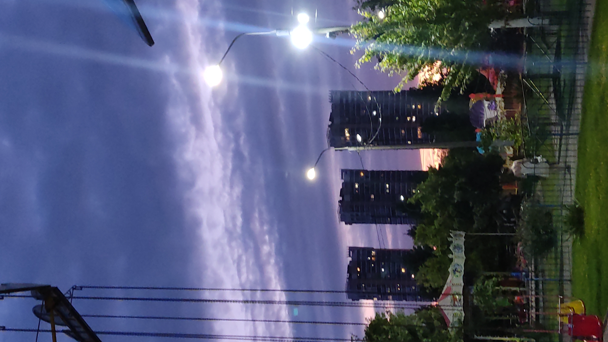 The image depicts a view of tall, modern buildings in an urban setting at dusk. The sky is a deep blue with streaks of purple and pink from the setting sun, and it is partially covered by gray clouds. In the foreground, there is a section of an amusement park, with a carousel visible on the left, and swings. A person in a light-colored dress is walking away from the camera towards the right side of the frame. Streetlights are illuminated, casting a bright glow and creating lens flares. The scene appears to be set in Krasnodar, Russia, given the architectural style of the buildings and the presence of an amusement park, which is common in residential areas.