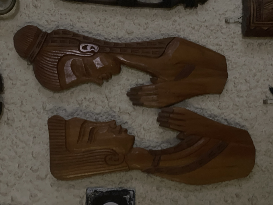 The image displays two decorative wooden relief carvings mounted on a textured, light-colored wall in Petah Tikva, Israel. The carvings depict the upper torsos of a male and a female figure, crafted from medium-brown wood, positioned facing each other in profile. Both figures exhibit elongated necks, closed eyes, prominent noses, and subtle smiles. The male figure, on the left, has vertically grooved hair styled flat on top. His hands are pressed together in a prayer-like gesture. The female figure, on the right, has vertically grooved hair gathered into a bun atop her head, with a visible earring or decorative element on her ear and white highlights on her face. Her hands mirror the male figure's, pressed together in a similar gesture. Both carvings feature carved lines suggesting clothing or adornment on their chests. The textured wall surface is visible behind and around the carvings. Partial views of other dark, decorative objects are visible on the periphery of the frame, specifically a dark rectangular object with a white circle to the far left and a darker, more intricate wooden carving with a metal hook at the bottom right.
