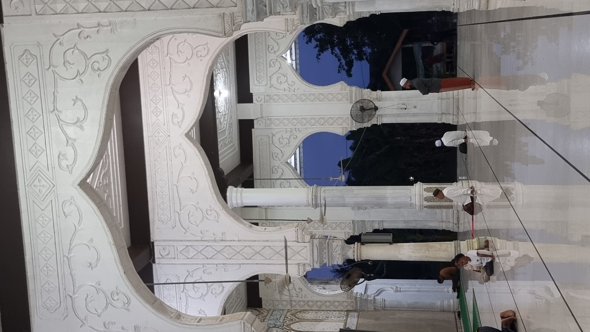The image shows the interior of a mosque in Meunasahlhok, Indonesia, characterized by ornate white Islamic architecture. Intricate carvings adorn the arches and columns, creating a sense of grandeur. Several people are present, engaged in prayer or sitting on the floor. In the foreground, a young boy in a white t-shirt is seated. Further back, several men in traditional Islamic attire are also seated or standing. The floor is highly reflective, mirroring the architectural elements and the figures. Through the archways, a dark blue sky and the silhouettes of trees are visible, suggesting it is either dusk or early evening. Electric fans are mounted on columns, indicating a tropical climate.