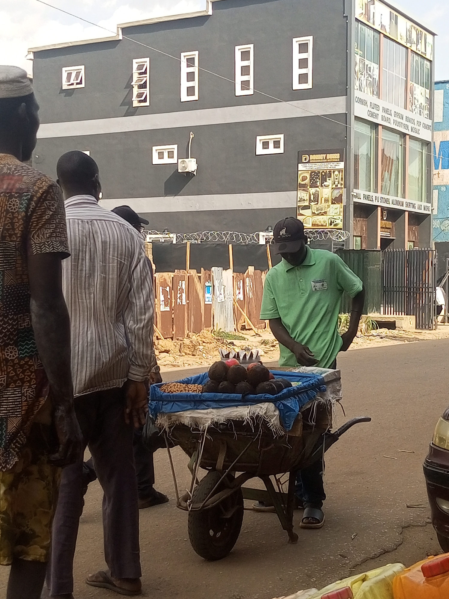 A street scene in Maiburiji, Nigeria shows several people standing around a vendor with a wheelbarrow full of produce, including peanuts and avocados. The vendor, wearing a green polo shirt and a black baseball cap, stands behind the cart. Behind the group are several buildings, including a gray building with white window frames and a taller building with a glass facade displaying various construction materials and advertisements. A fence topped with barbed wire is also visible in the background. A car's headlight is visible on the right side of the image.