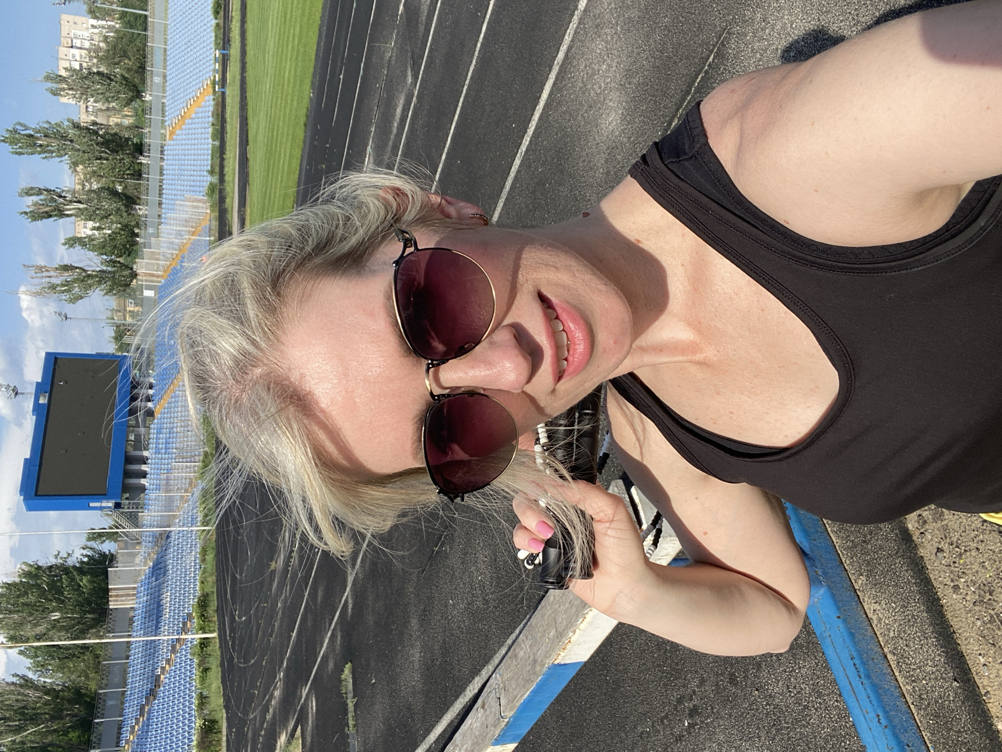 A woman takes a selfie on a running track at a stadium. The stadium has blue seating, a green lawn, and a blue scoreboard. The woman is wearing sunglasses and a black tank top. She is holding a small, black object in her hand. The location is in the city of Unknown, Unknown.