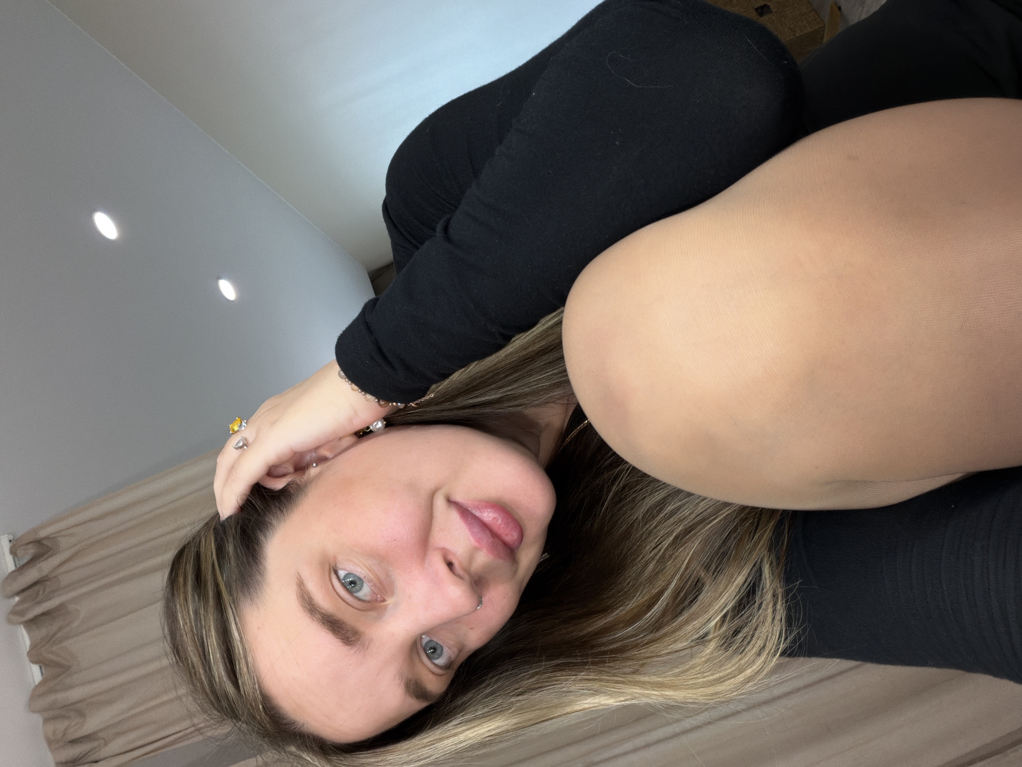 A woman with fair skin and blue eyes is looking at the camera. She has blonde hair with darker roots and is wearing a black long-sleeved shirt. Her right hand is touching her hair, and she is wearing jewelry on her fingers and wrist. In the background, there is a light beige curtain and a white wall with recessed lighting. The location is not discernible from the image.