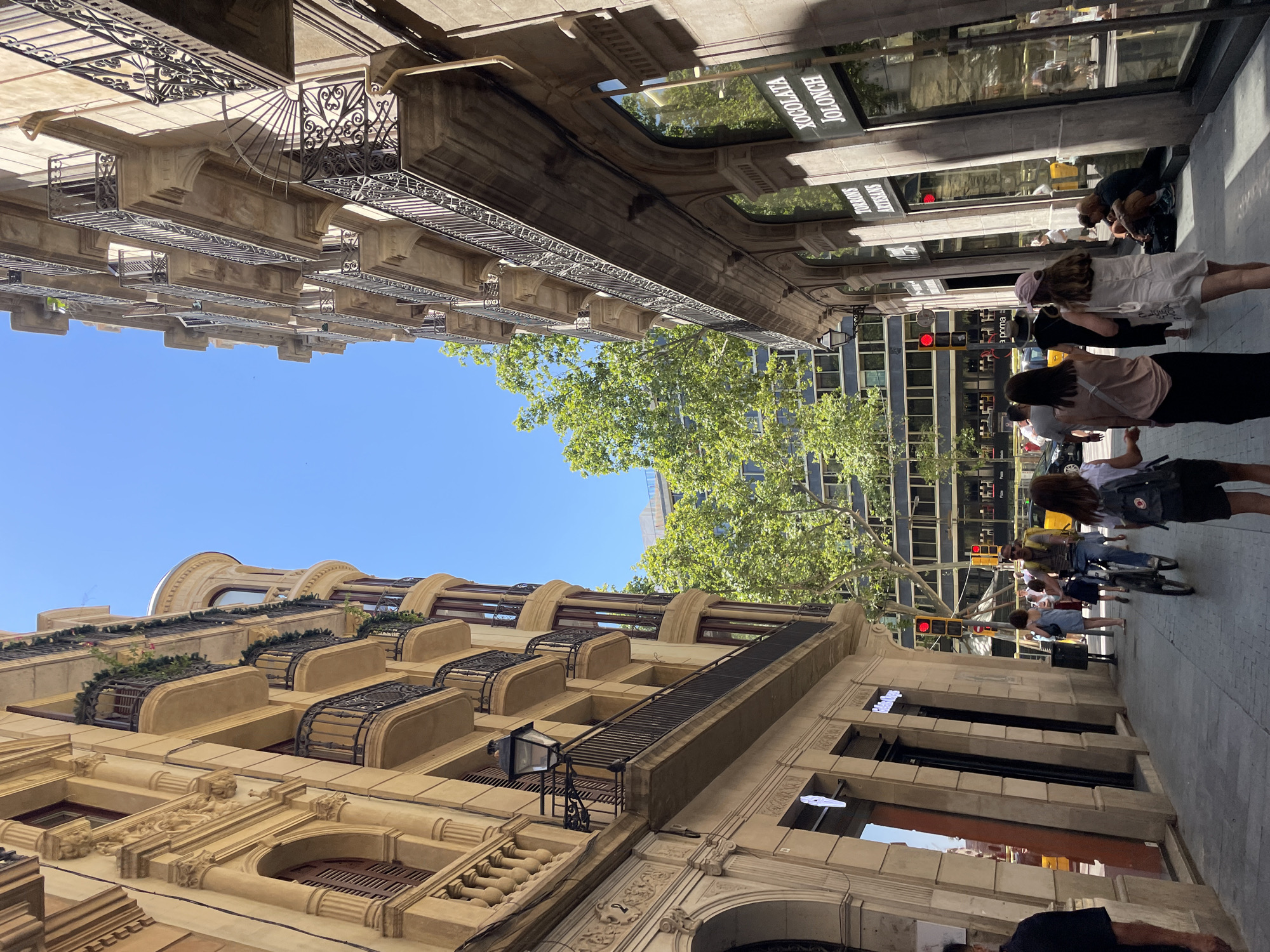 This is a low-angle, wide shot of a bustling street in Barcelona, Spain, on a sunny day. The perspective is tilted, giving the impression that the buildings are leaning. In the foreground, ornate, tan-colored buildings with intricate stonework and wrought-iron balconies rise upwards. Some balconies are decorated with greenery.
The street is filled with people walking and cycling. A few individuals are visible in the midground, some appearing to be tourists. A yellow taxi is partially visible on the right side of the frame. In the background, modern glass-fronted buildings can be seen interspersed with trees. Traffic lights are visible, one showing red.
Visible text includes "JOLLOCHATA" and "JOLLOSHO" on signs in shop windows. The clear blue sky and bright sunlight suggest it is midday. The overall atmosphere appears lively and urban, typical of a busy city street.