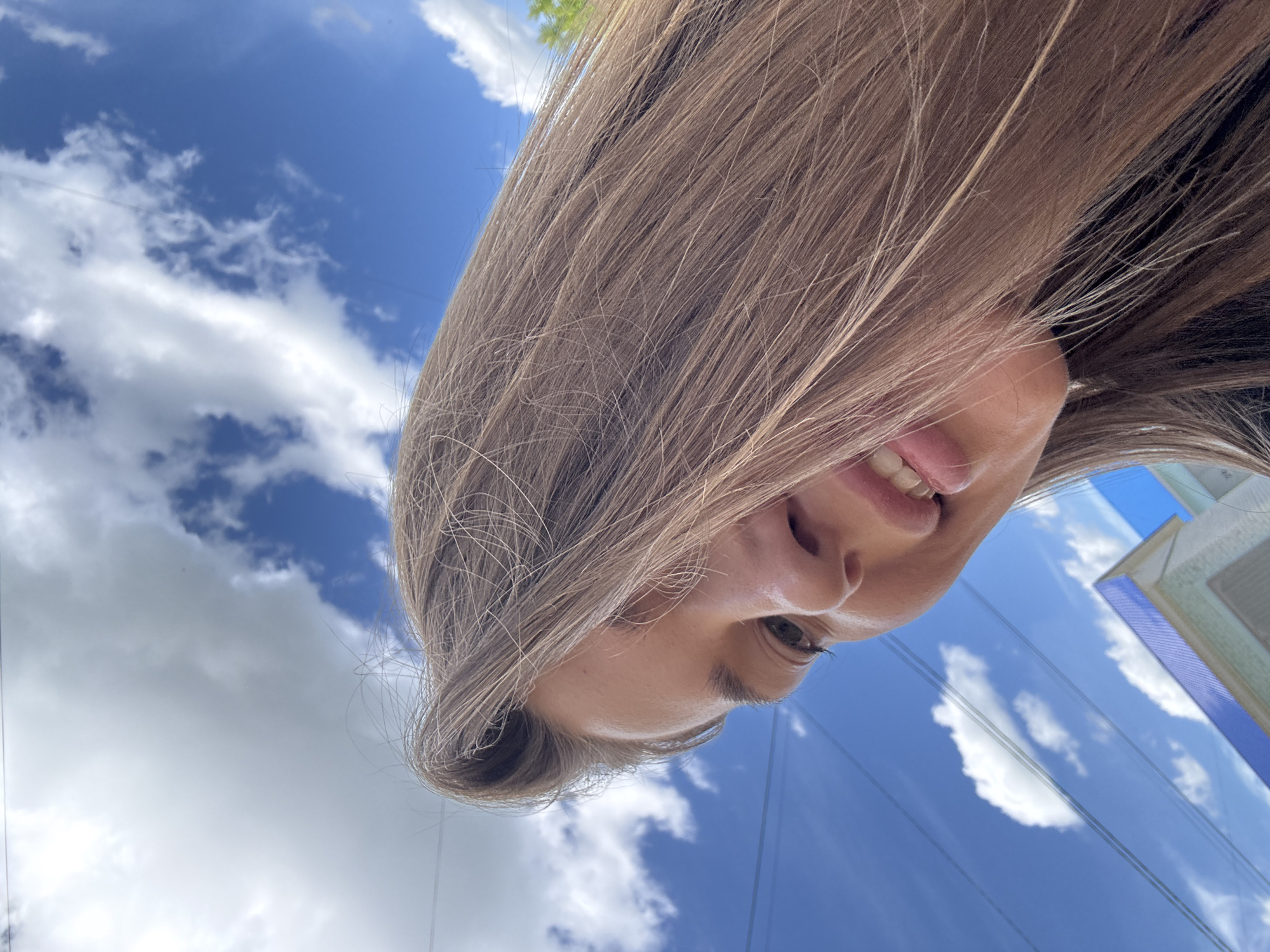 In the city of Chernushka, Russia, an individual is captured from a low angle, looking upwards towards a bright blue sky dotted with white, fluffy clouds. The subject's long, brown hair fills a significant portion of the frame, partially obscuring their face. Their eyes are open and looking slightly downwards. Their mouth is slightly open, revealing teeth and what appears to be a tongue. The lower portion of a building with a blue roof is visible in the background, as are power lines stretching across the sky. The overall lighting suggests it is daytime.