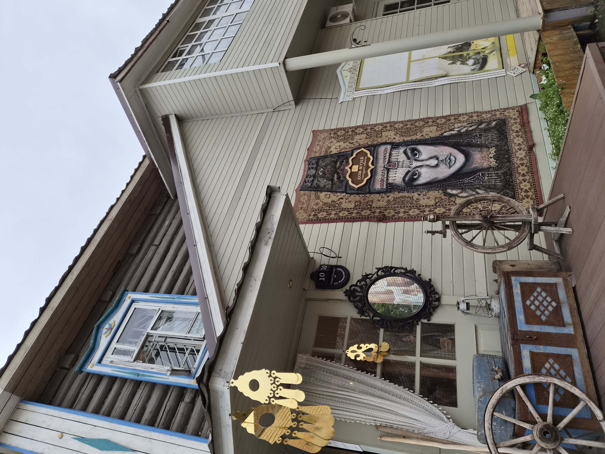 This image shows the exterior of a building, likely a museum or cultural center, in Kazan, Russia. The building features a combination of traditional wooden log construction on the upper level and lighter colored siding on the lower levels.
Several decorative elements adorn the facade. Two large, ornate gold-colored earrings hang from the porch roof. Below them, a white lace curtain covers a portion of a window. A black, ornate mirror hangs on the wall, reflecting a blurred image. A sign with the numbers "10 10" is visible near the mirror.
A large tapestry featuring a portrait of a woman in traditional attire is hung on the lower wall. To the right of this tapestry, another decorative panel with floral motifs is displayed.
In the foreground, a wooden porch is decorated with several antique-looking items. A large wooden wheel is positioned on the left. A weathered wooden chest with blue and brown geometric patterns sits behind it, and a vintage lantern is placed on top of the chest. Another wooden spinning wheel is located on the right side of the porch. A flower box filled with green plants and white flowers is visible at the base of the building.
The sky is overcast, suggesting a cloudy day. The overall impression is one of a culturally rich and artistically decorated location.